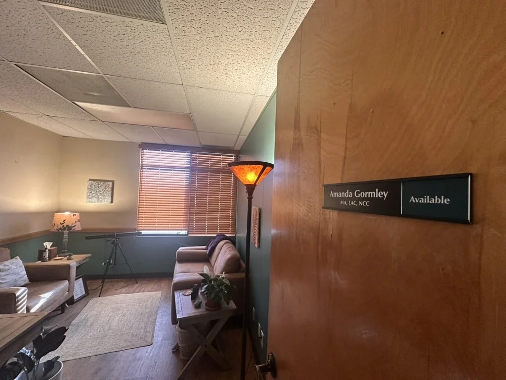 Entrance to therapy room with a door labeled Amanda Gormley, available, and showing a sofa, armchair, side table, potted plant, lamp, window with blinds, EMDR machine, and wall decor.
