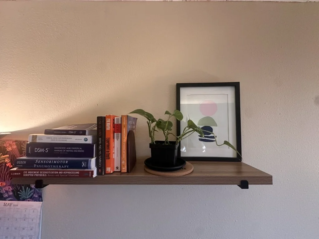 A wooden shelf in a therapist's office holding a stack of books, a framed abstract art print, and a potted plant.