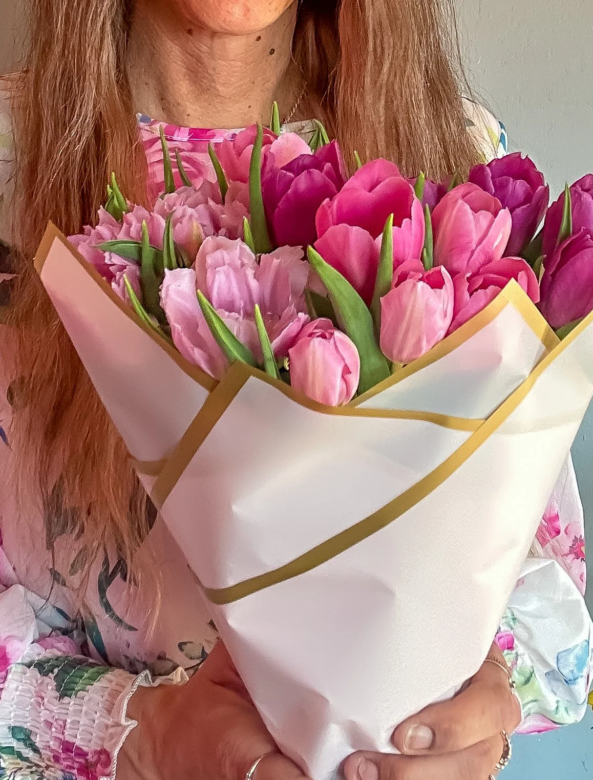 Person holding a bouquet of pink, purple, and fuchsia tulips wrapped in white and gold paper.