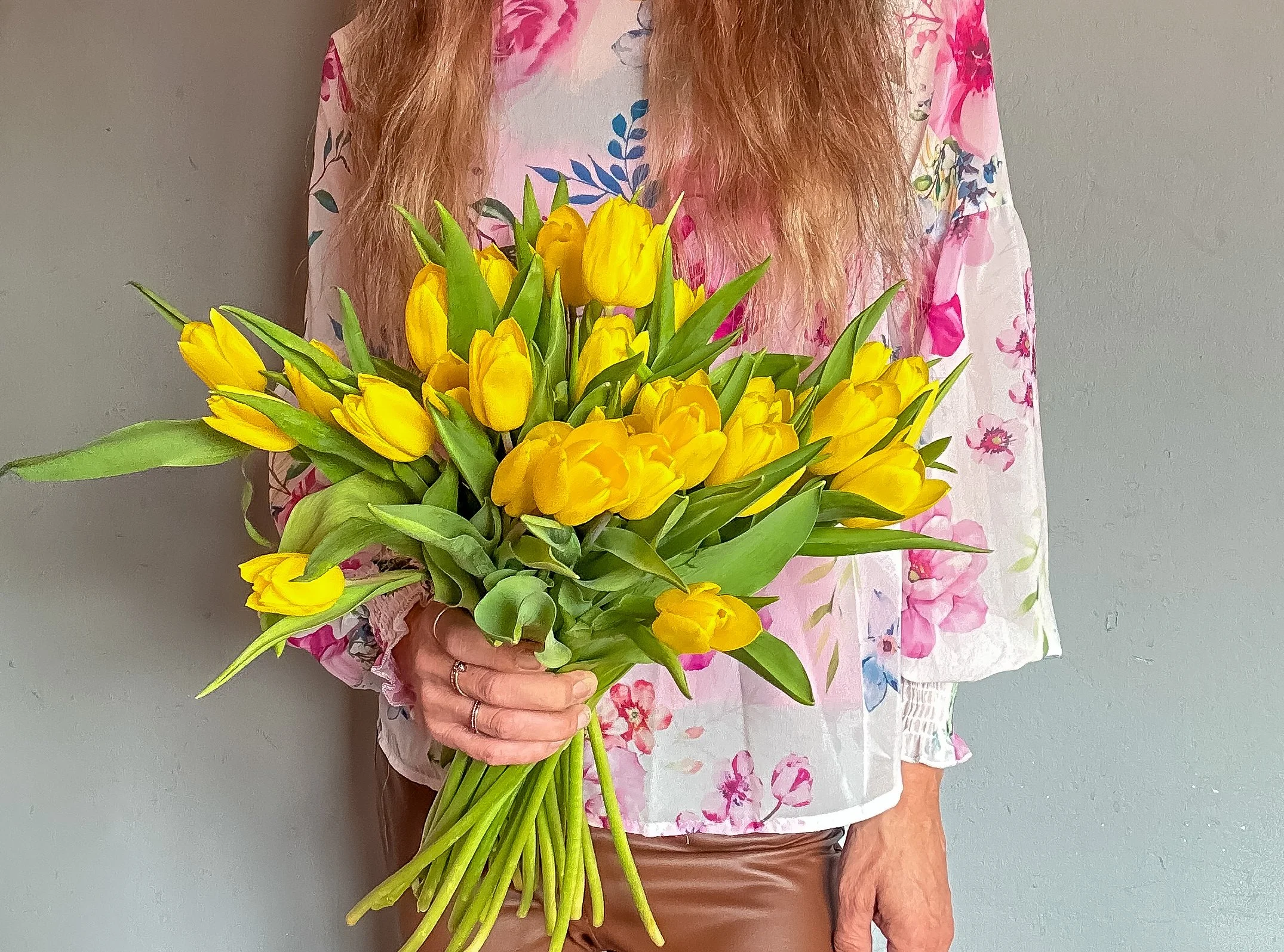 Person holding a large bouquet of yellow tulips, wearing a floral pink and white blouse and brown pants, against a plain gray wall.