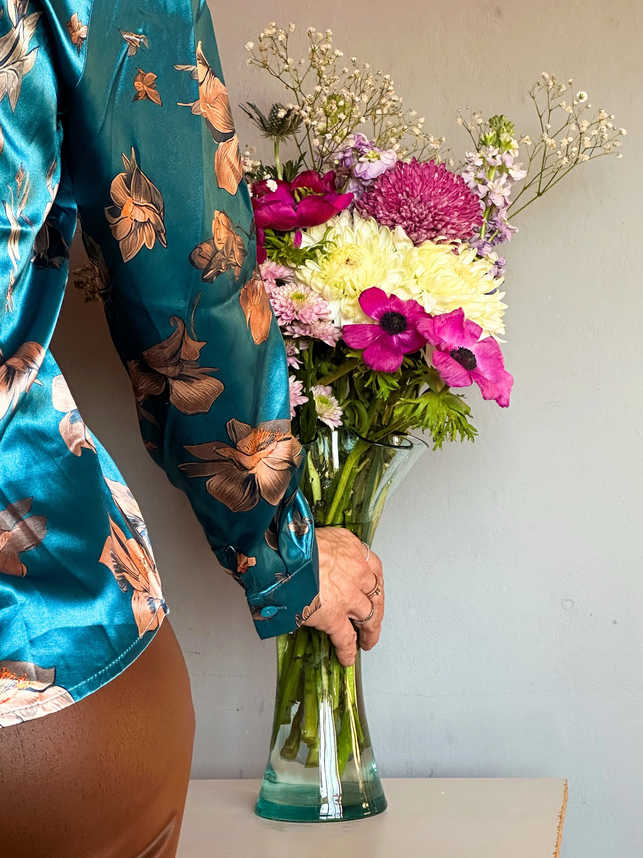 A person dressed in a teal satin blouse with floral print holding a large glass vase of mixed colorful flowers, including pink, yellow, and white blooms, against a plain wall.