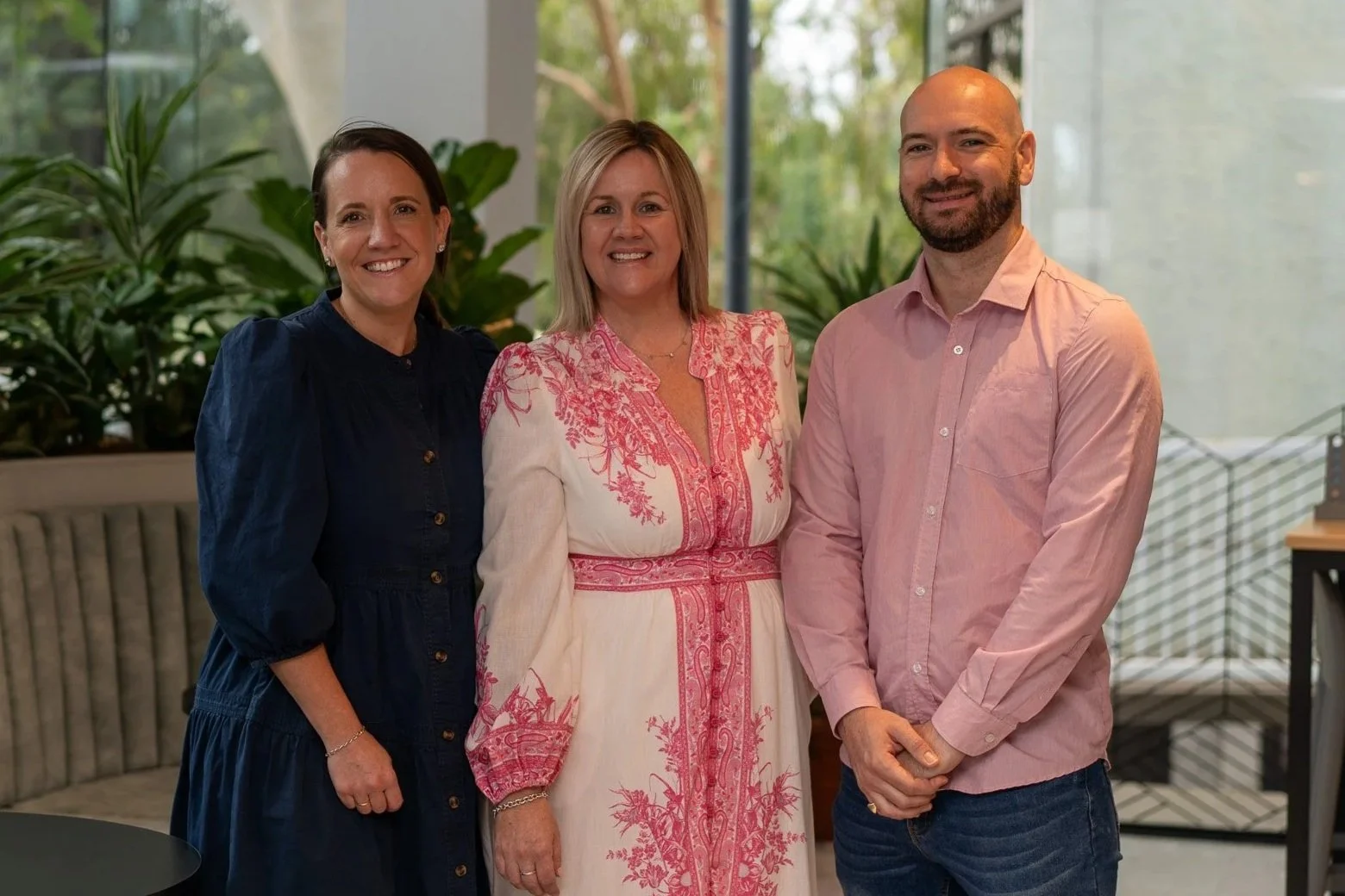 Three adults standing in an office or indoor space with large windows and green plants behind them. They are smiling at the camera.