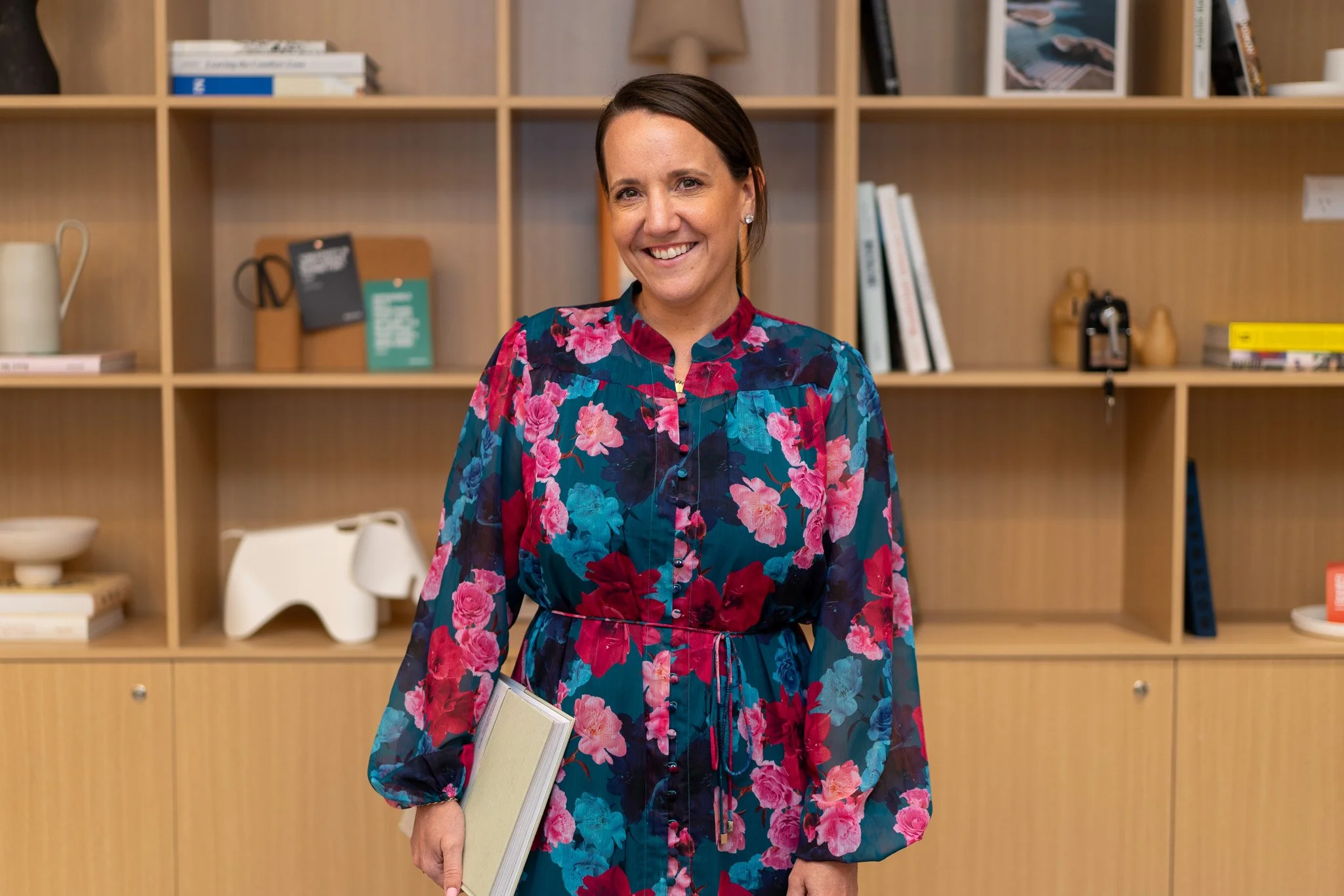 A smiling woman wearing a floral dress standing in front of a wooden shelf filled with books and decorative items, holding a notebook.