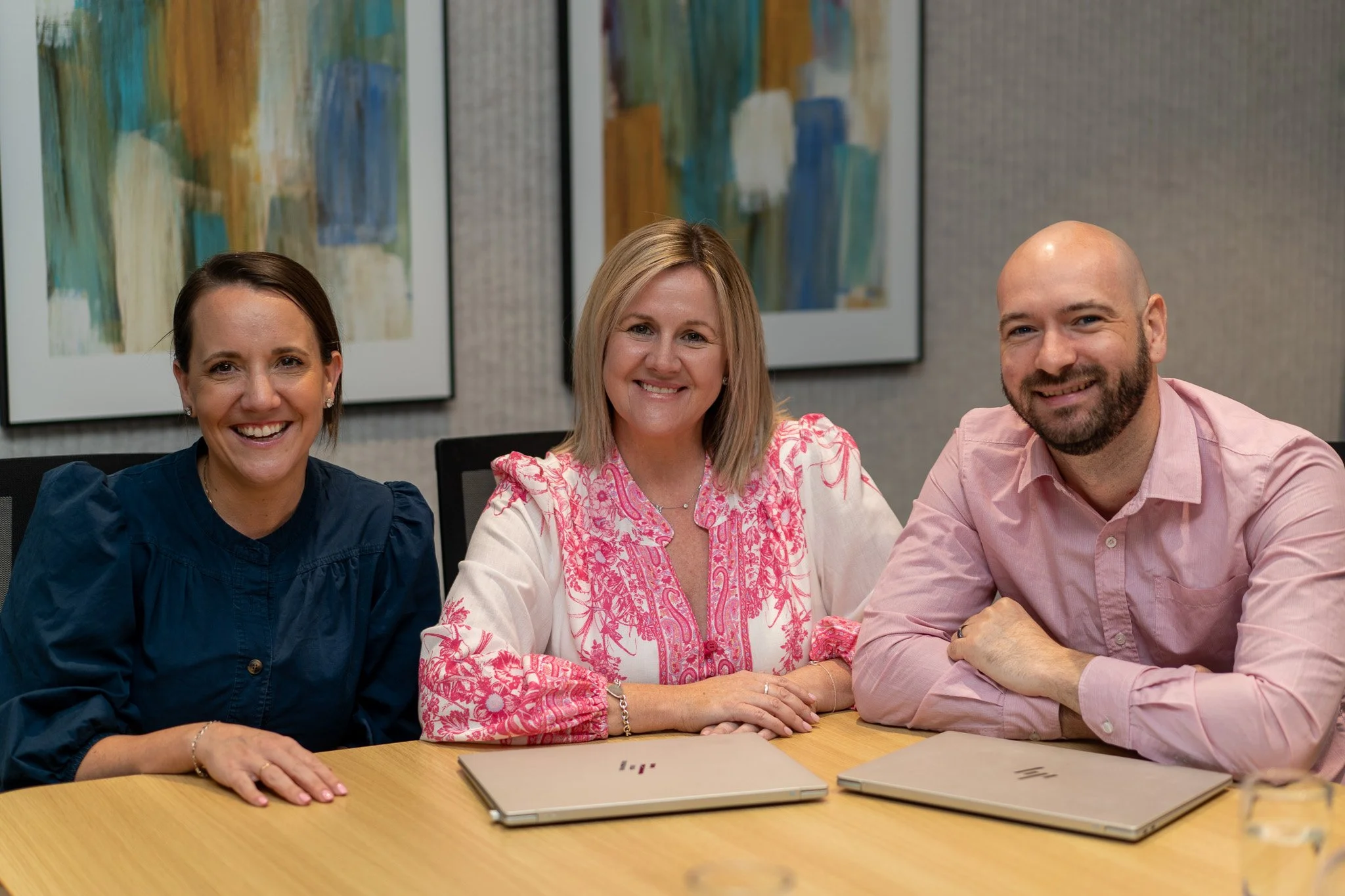 Three people sitting at a table, smiling, with laptops in front of them, in a conference room with abstract art on the wall.