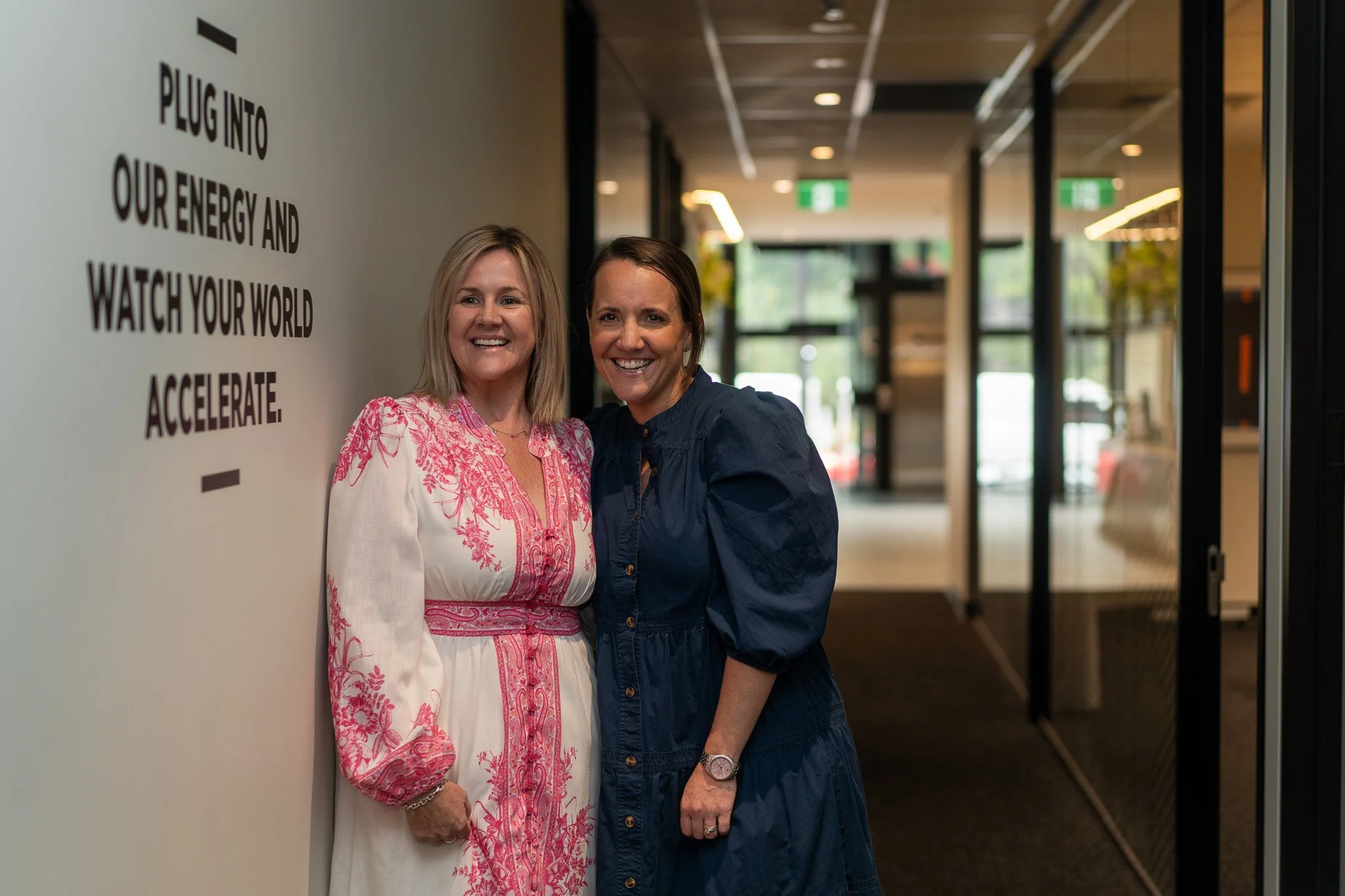 Two women smiling and standing close together indoors near a wall with a motivational quote, in a modern building with glass doors and windows in the background.