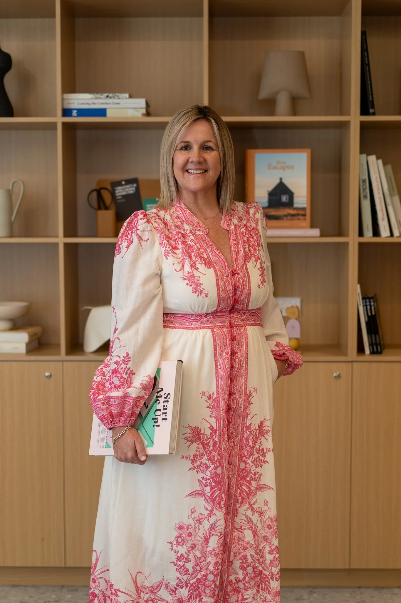 A woman in a white and pink embroidered dress holding a book titled 'Start Your Move' with a bookshelf in the background.