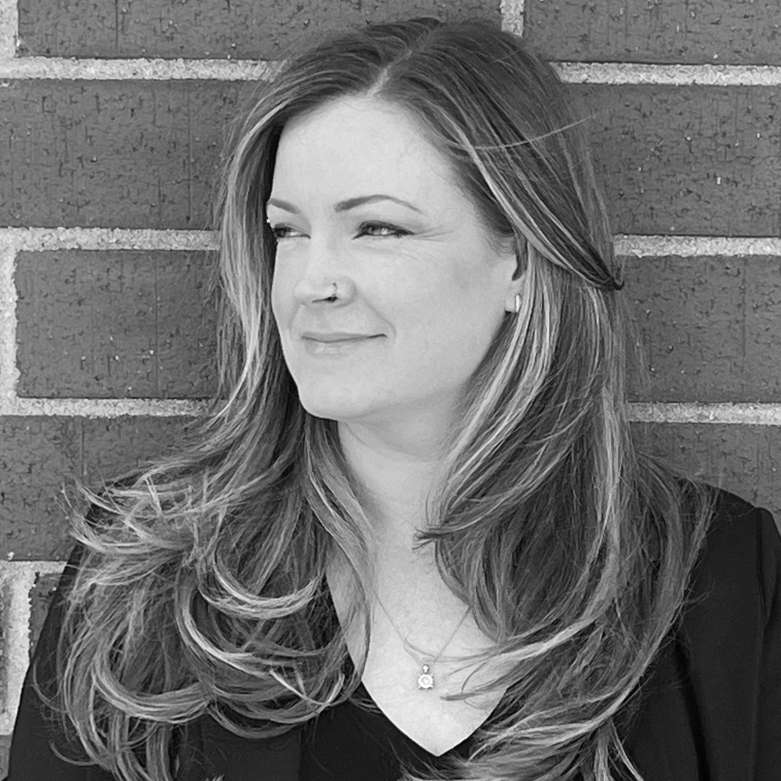 Black and white portrait of a woman with long, wavy hair, wearing a necklace and a dark top, standing against a brick wall, looking to the side with a slight smile.