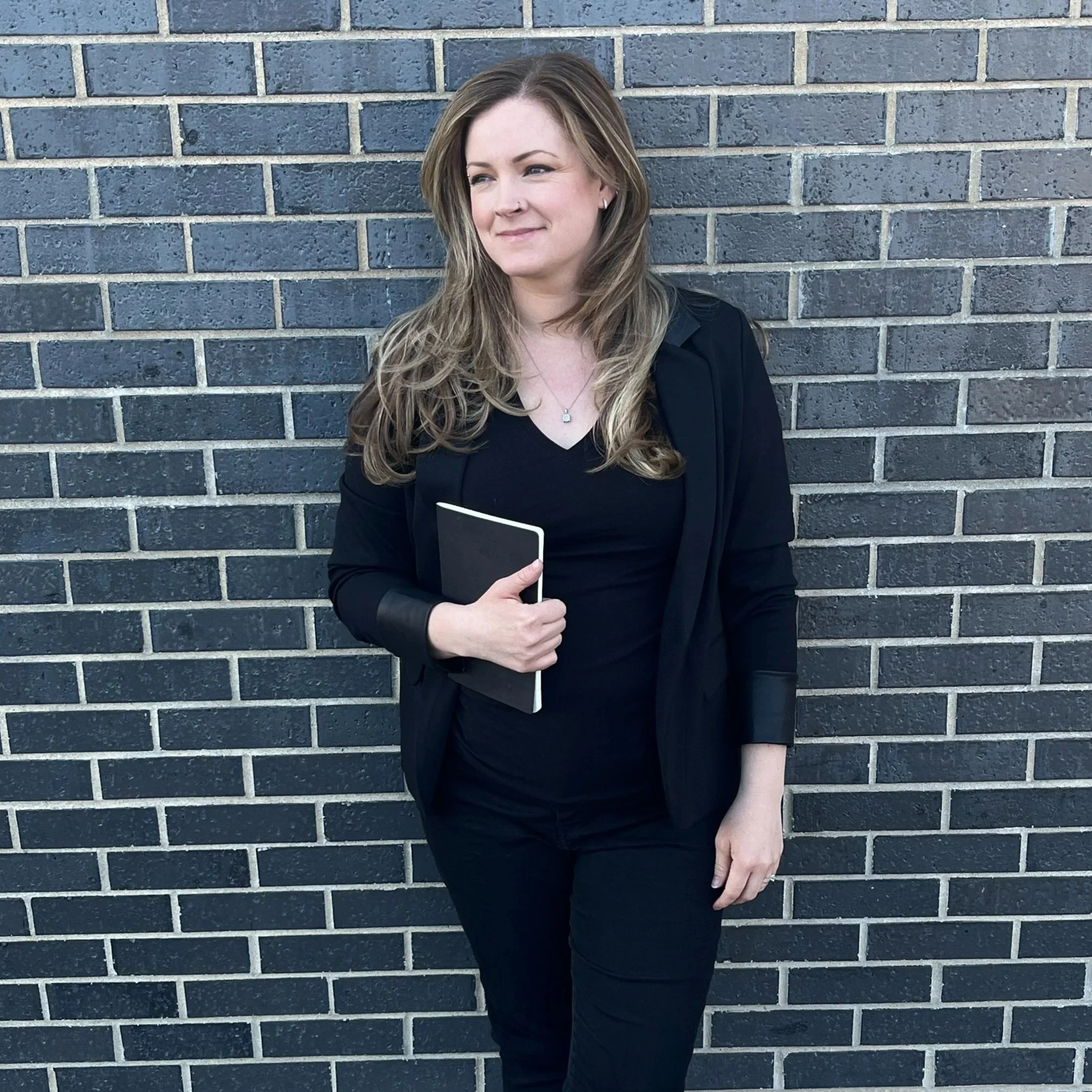 A woman with long wavy hair wearing a black blazer, black top, and black pants, standing against a brick wall, holding a black notebook.