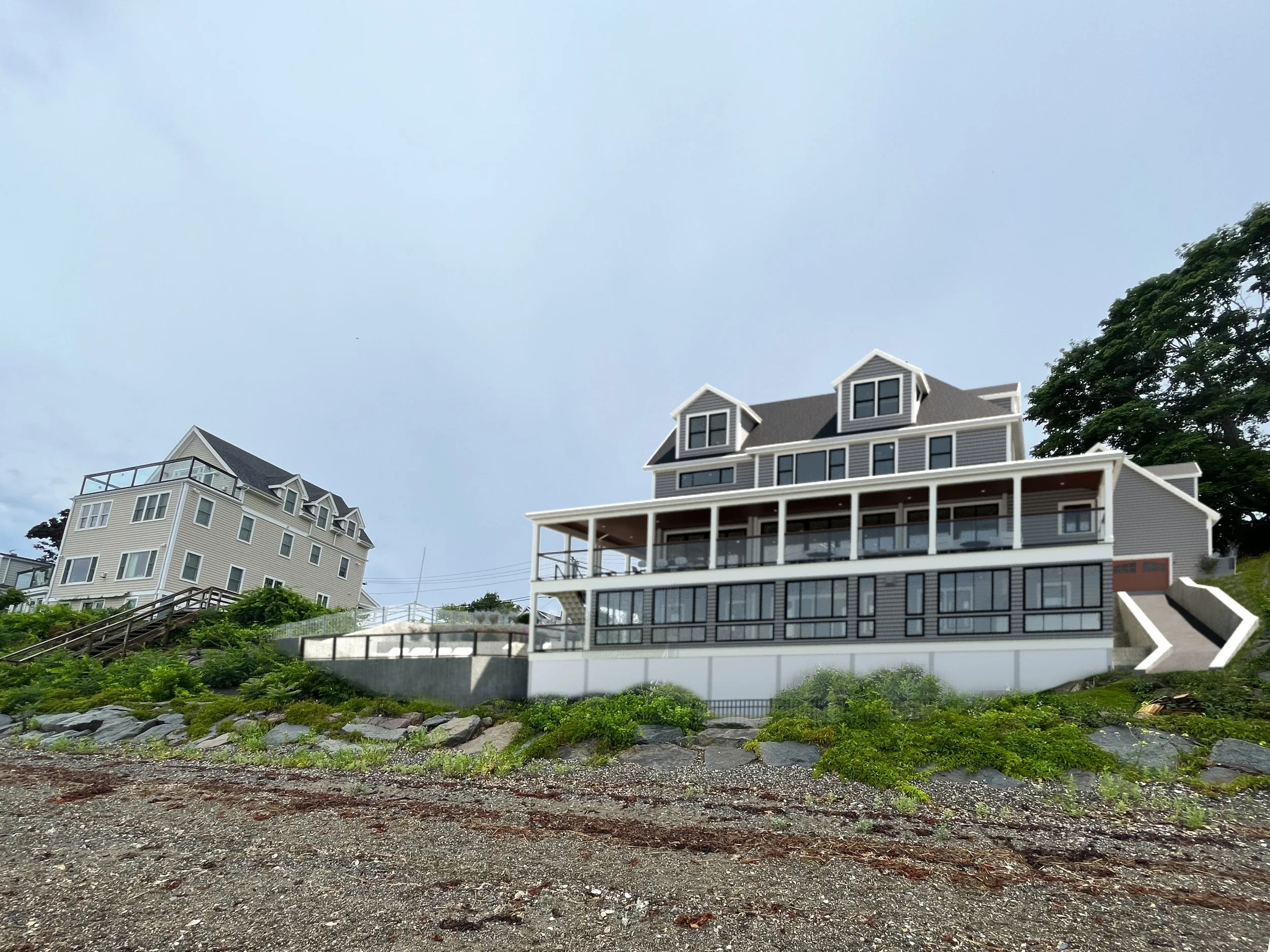 Large multi-story house on a hill with a view of the ocean, light grey siding, multiple gabled roofs, large balcony, and surrounding greenery.
