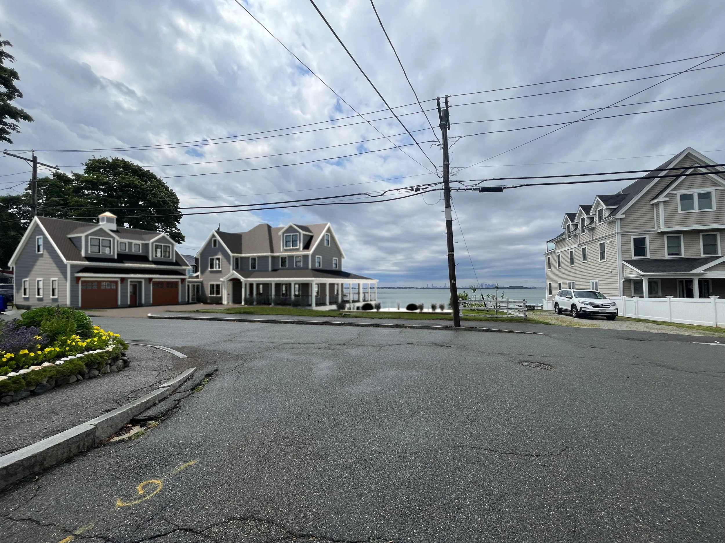 Residential neighborhood near waterfront with three houses, a boat launch, and a car parked on the street against a cloudy sky.