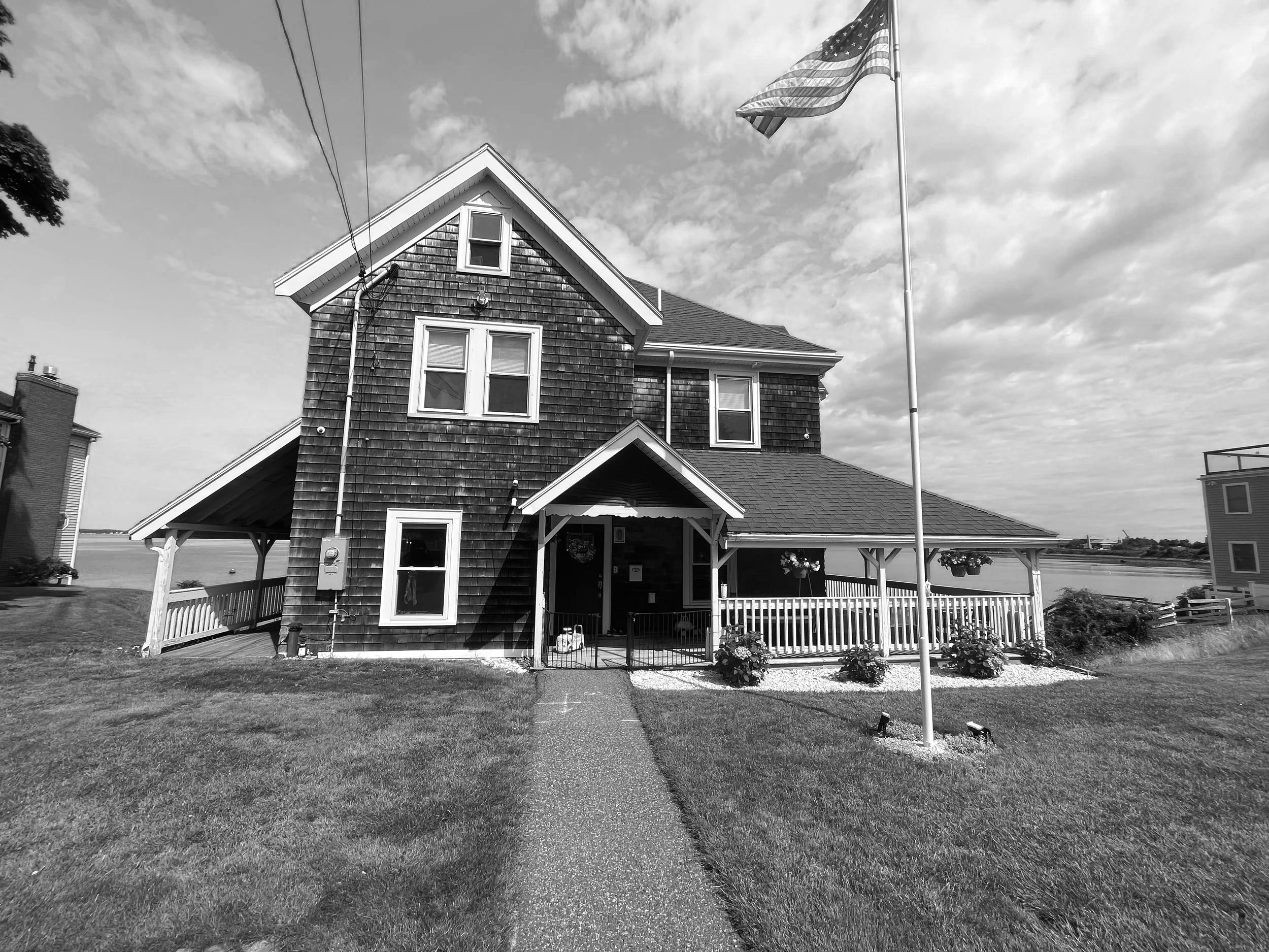 Black and white photo of a two-story house with a porch, American flag, and well-kept lawn, with a body of water in the background.