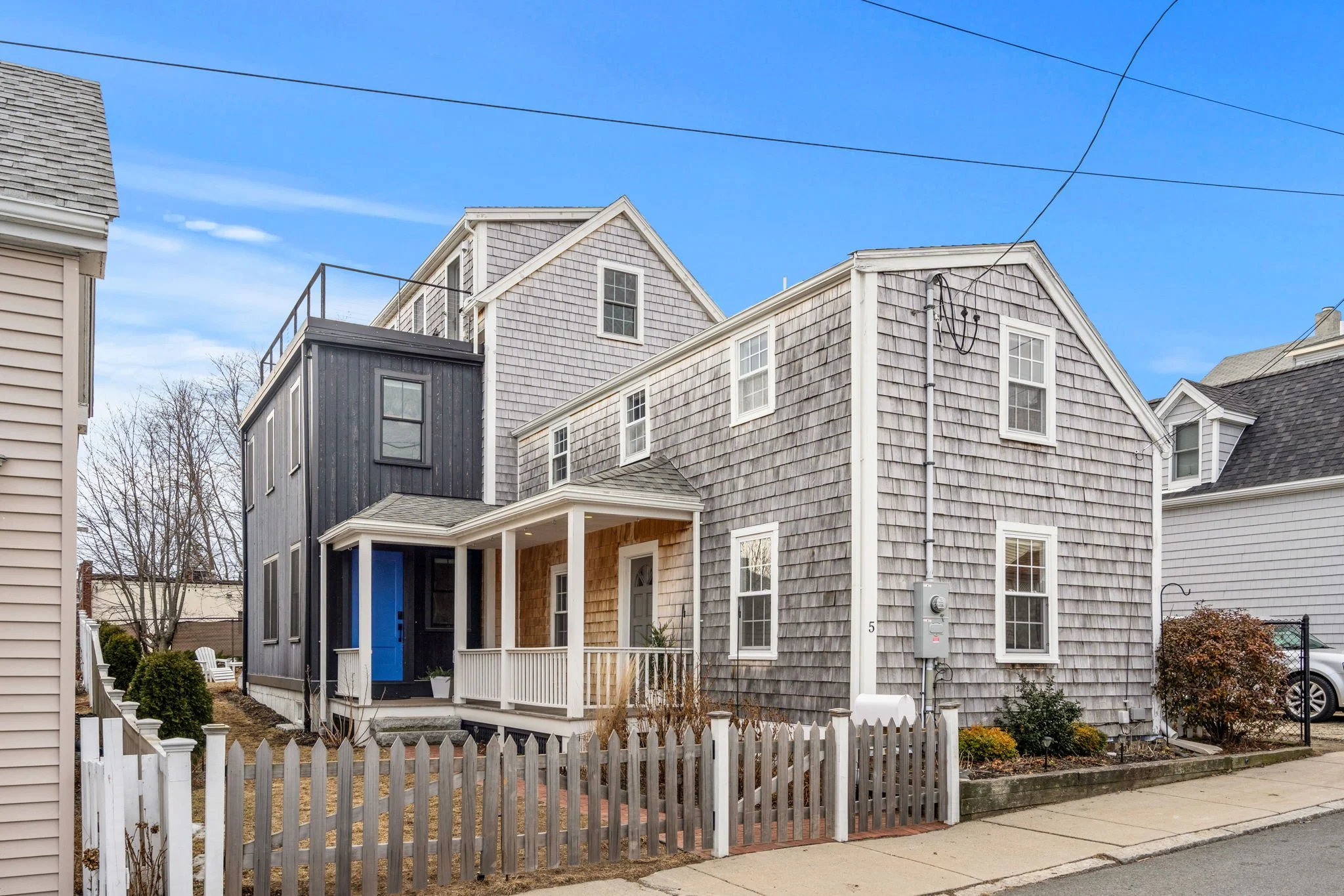 A modern multi-story house with gray shingle siding, a small front porch, and a white picket fence, located in a suburban neighborhood under a clear blue sky.