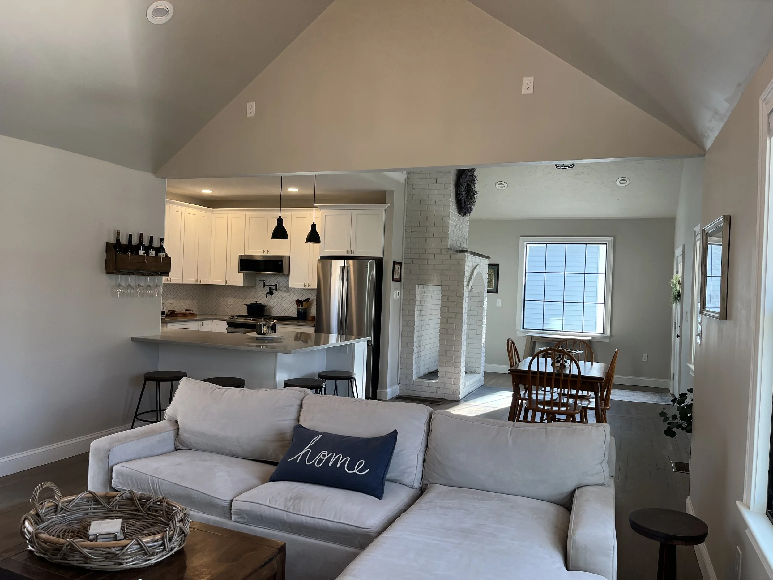 Open-concept living room with white sofa, navy throw pillow with 'home' label, wicker basket, view of kitchen with white cabinets, stainless steel refrigerator, black pendant lights, dining area with wooden table and chairs, front door with window, and fireplace with white brick surround.