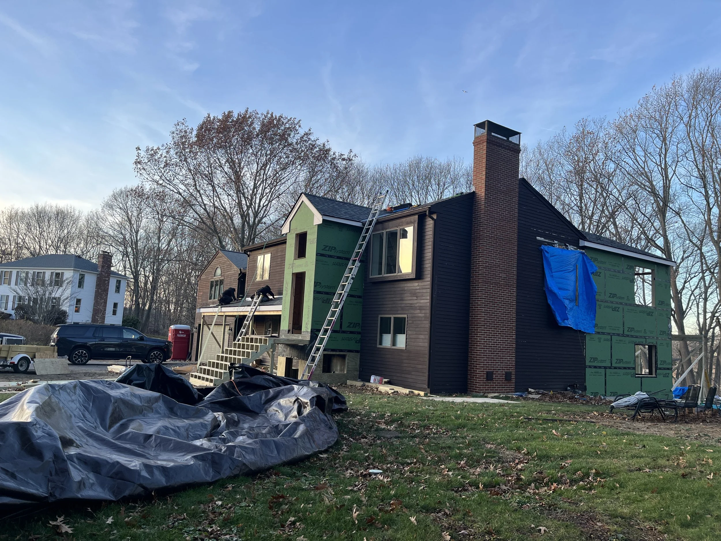 A house under construction with siding partially installed, ladders leaning against it, and construction materials on the ground. The sky is clear and the surrounding trees are leafless.