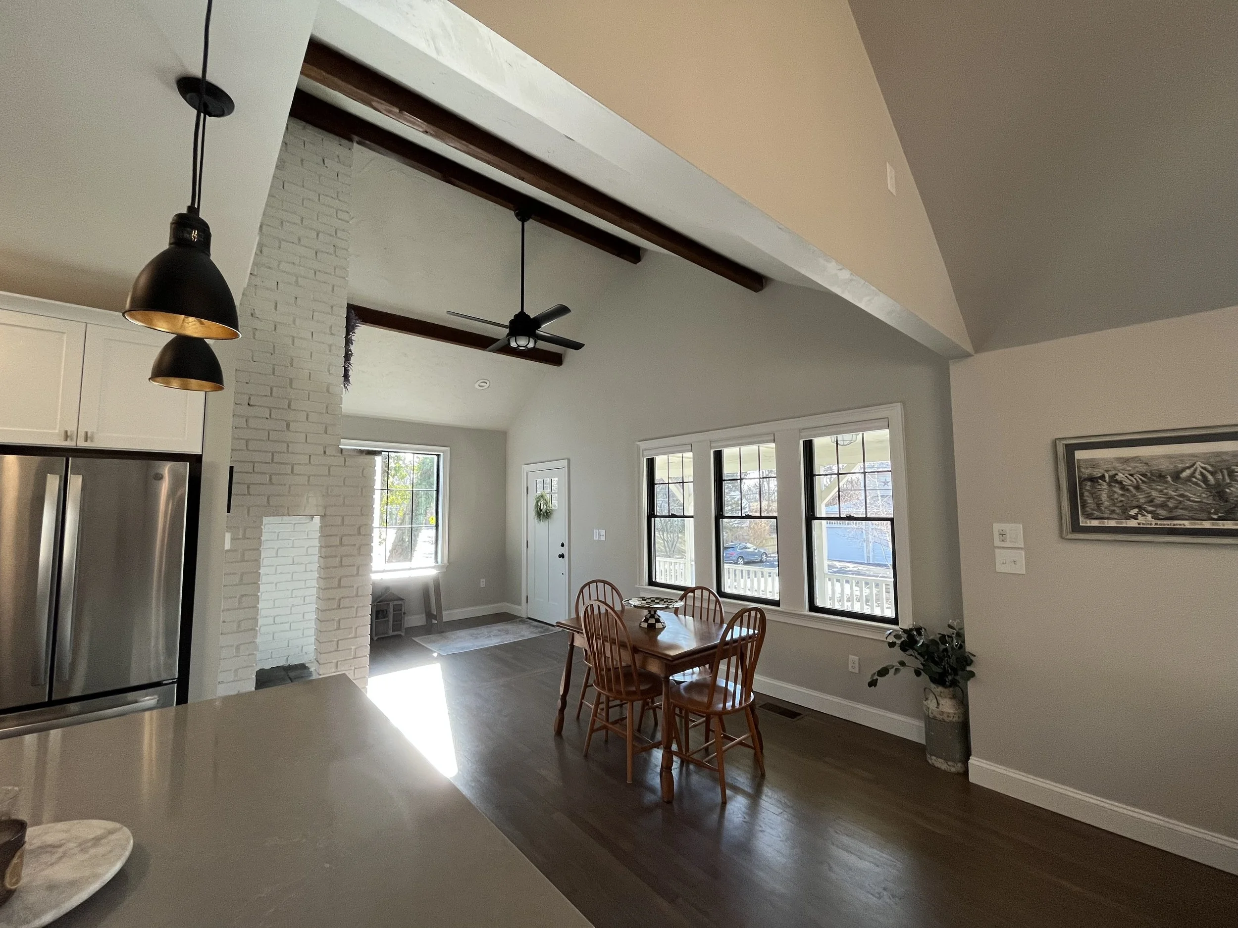 Interior of a living room and dining area with large windows, a white brick fireplace, wooden dining table with four chairs, plants, and a high ceiling with beams and ceiling fans.