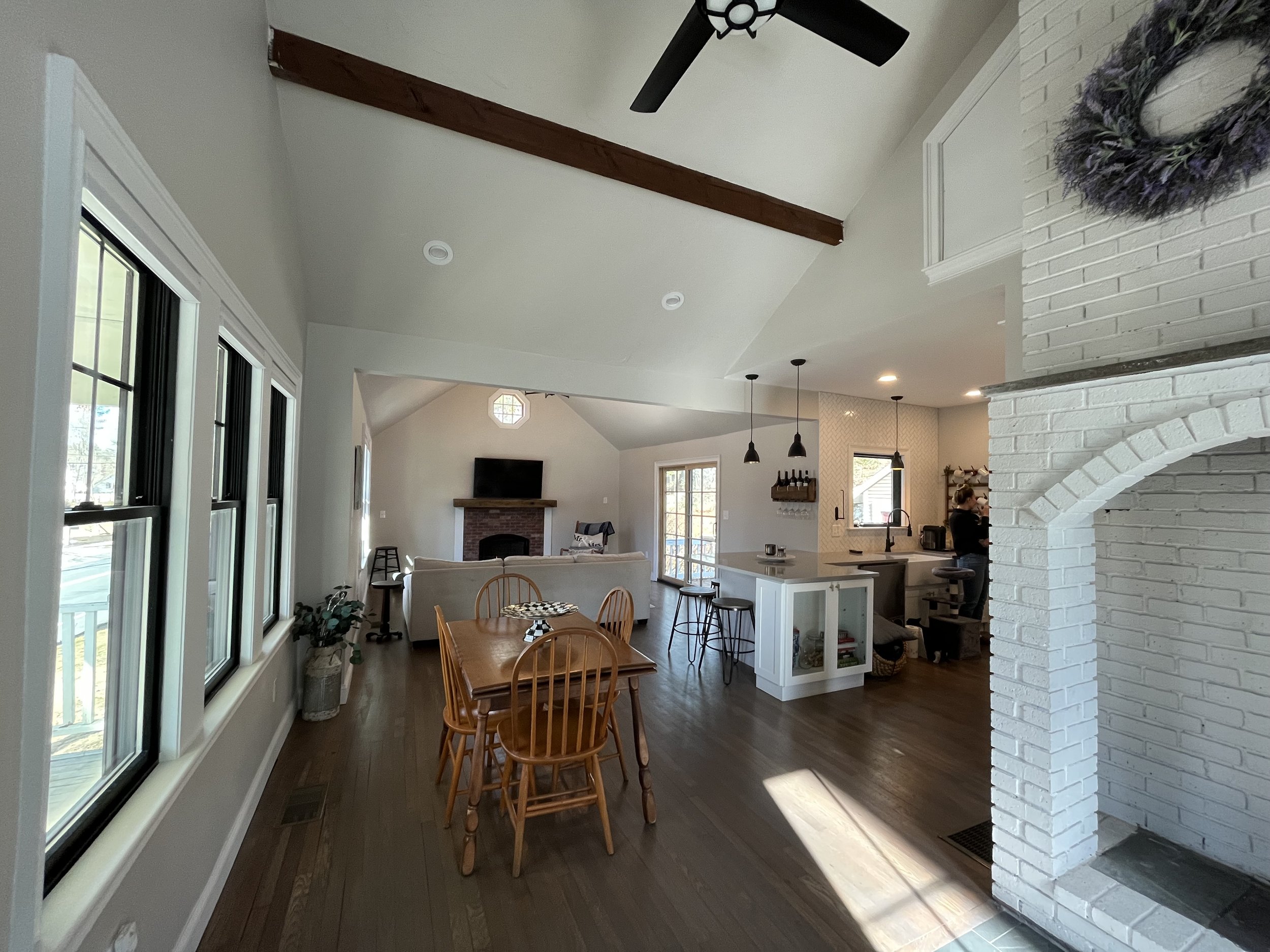Open-concept living and dining area with white walls, large windows, hardwood floors, a white brick fireplace, a wooden dining table with chairs, a kitchen with a white island, black pendant lights, and a woman preparing food.