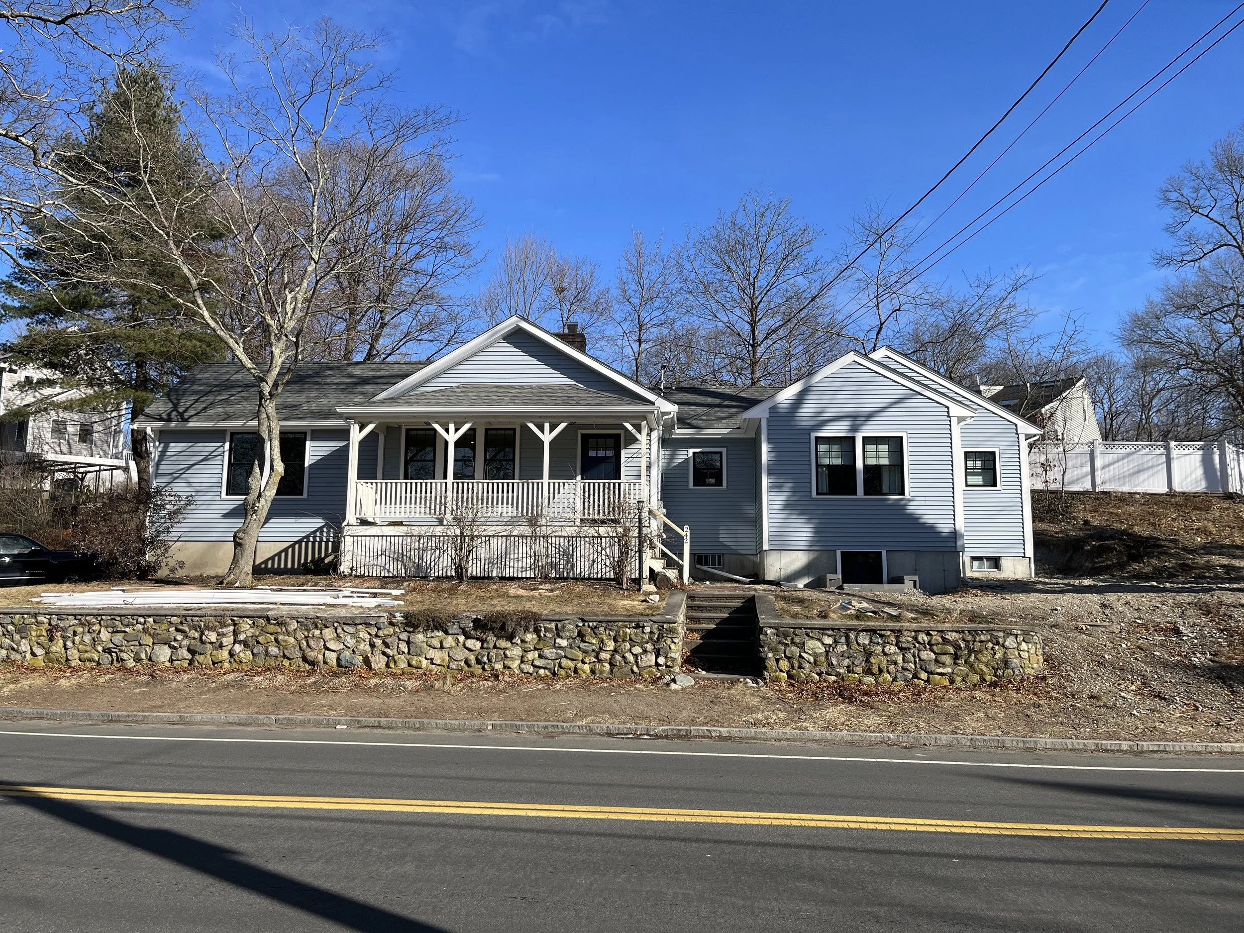 A house with blue siding, a porch with white railings, and several windows, set on a slightly elevated area with stone retaining walls and stairs leading up to the house. There are bare trees and a clear blue sky in the background.