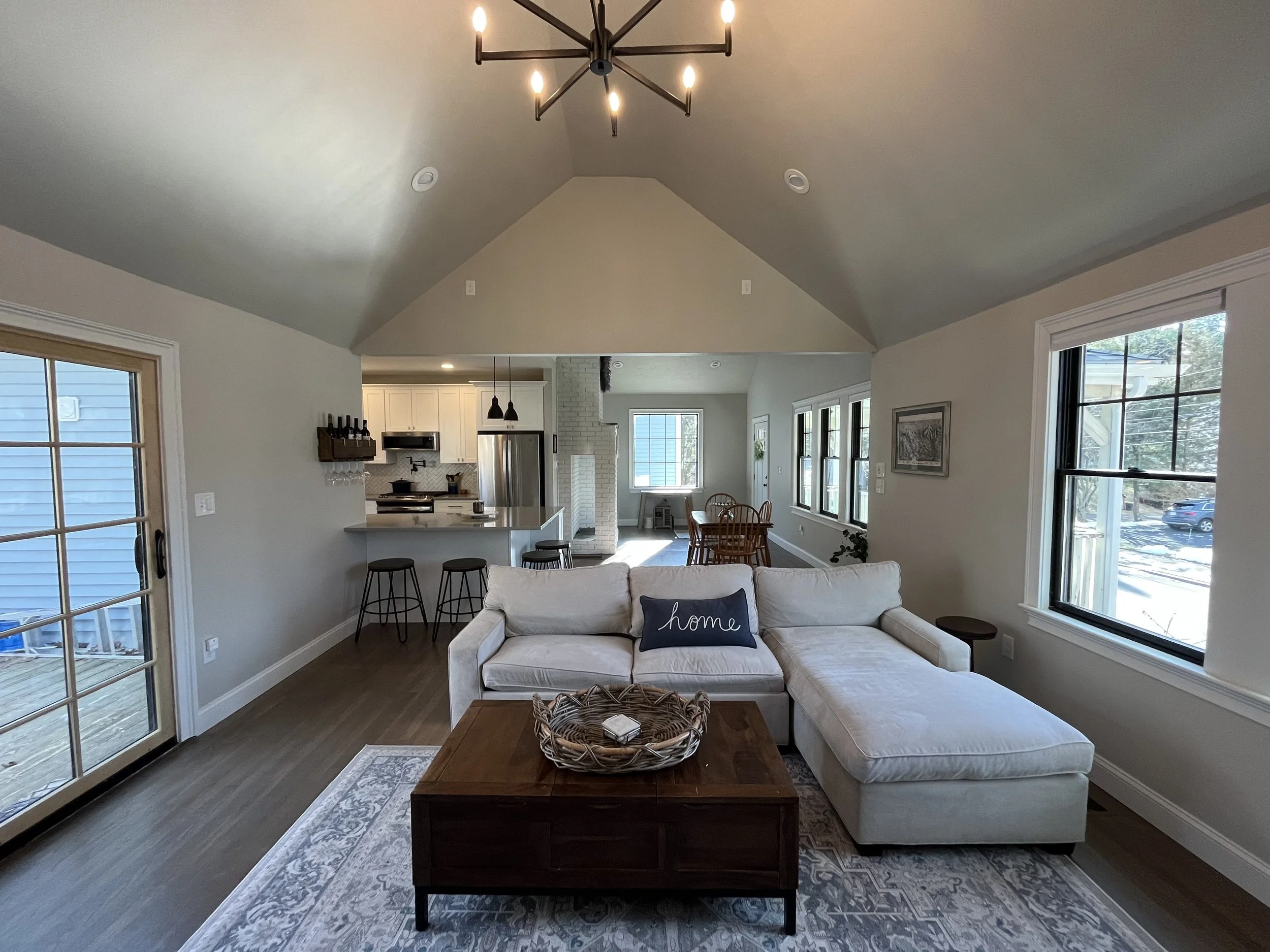 Living room with a white sectional sofa, a wooden coffee table with a basket, large windows, and a view of the kitchen and dining area.
