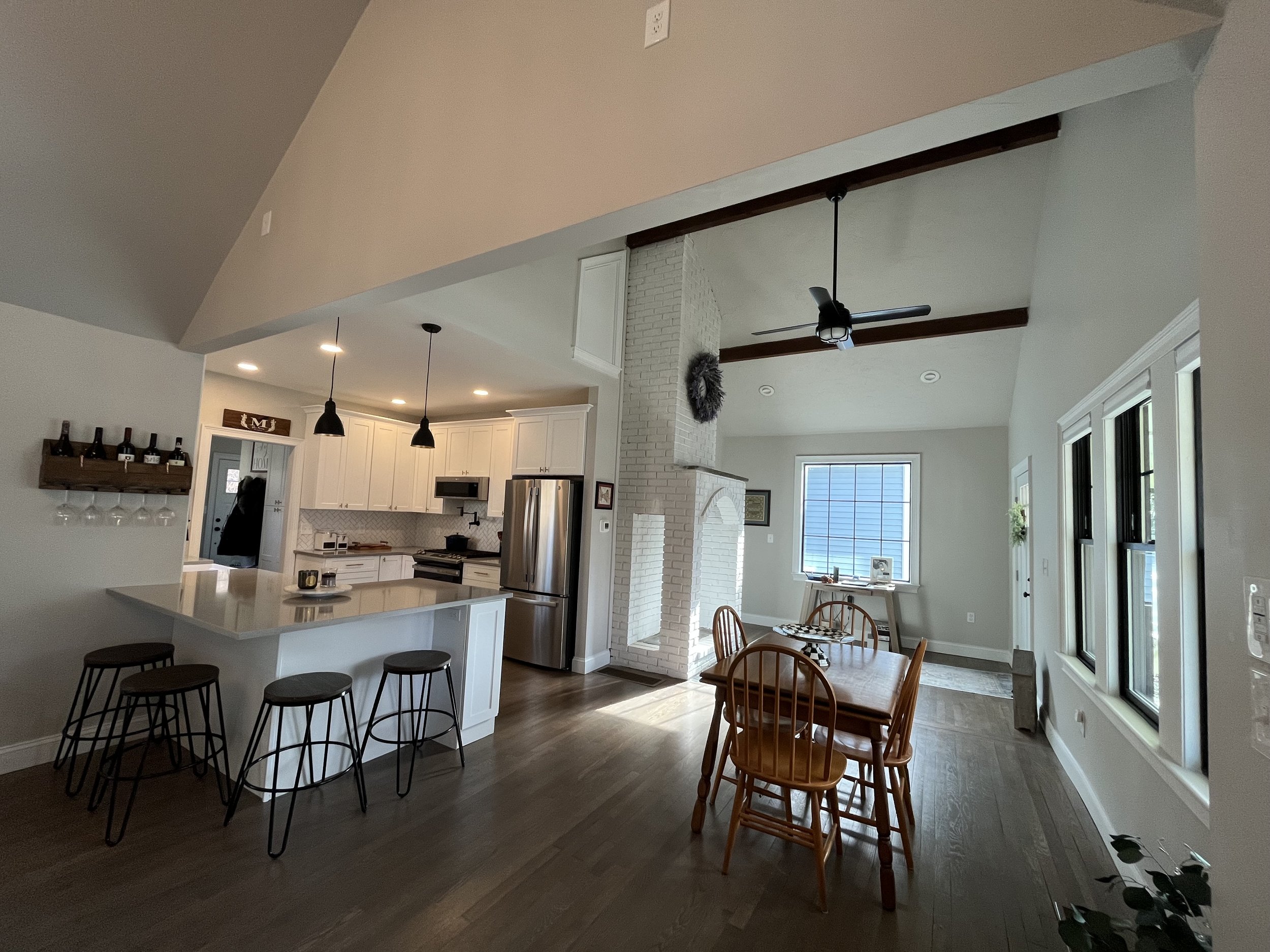 Open-concept kitchen and dining area with white cabinets, stainless steel refrigerator, island with bar stools, and a dining table with chairs. Large windows provide natural light. A brick fireplace is visible near the dining area.