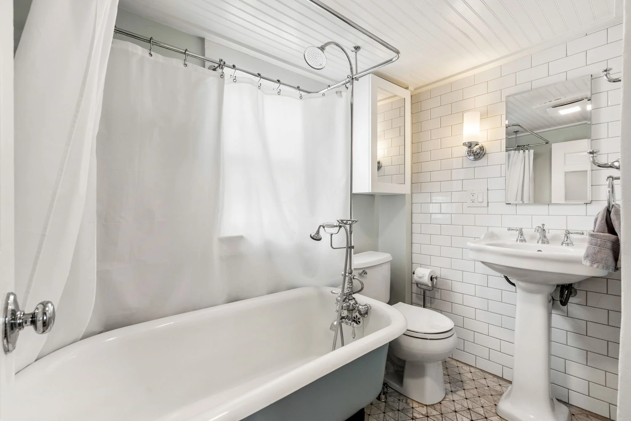 A bathroom with a white shower curtain, vintage bathtub with fixtures, white toilet, pedestal sink, and a mirror on a white tiled wall with wall-mounted light fixtures.