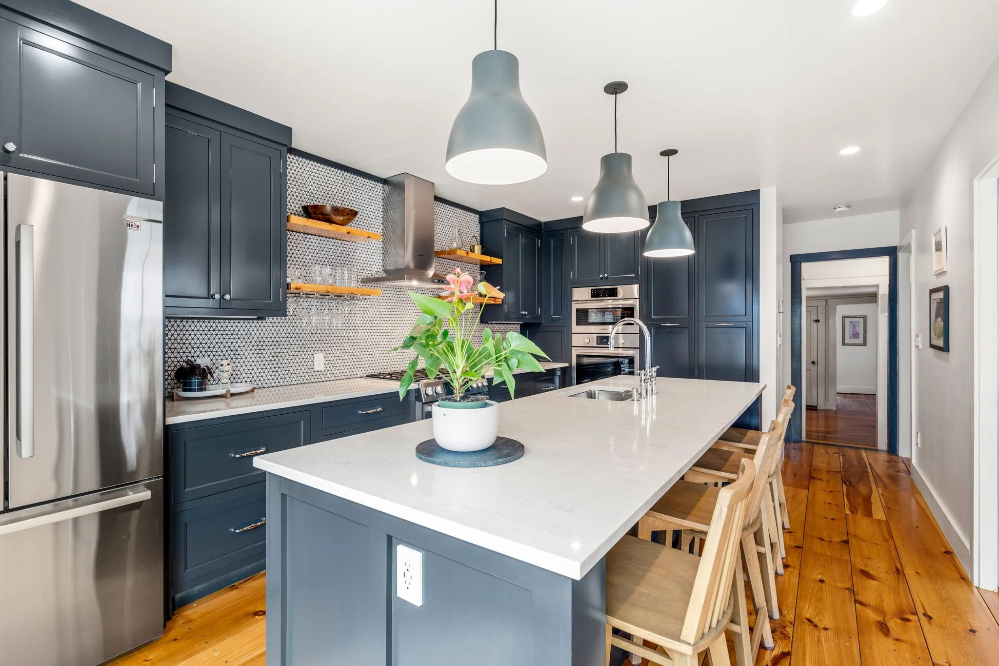 Modern kitchen with navy blue cabinets, a white island with a potted plant, stainless steel appliances, a black and white geometric backsplash, pendant lighting, and wooden stools.