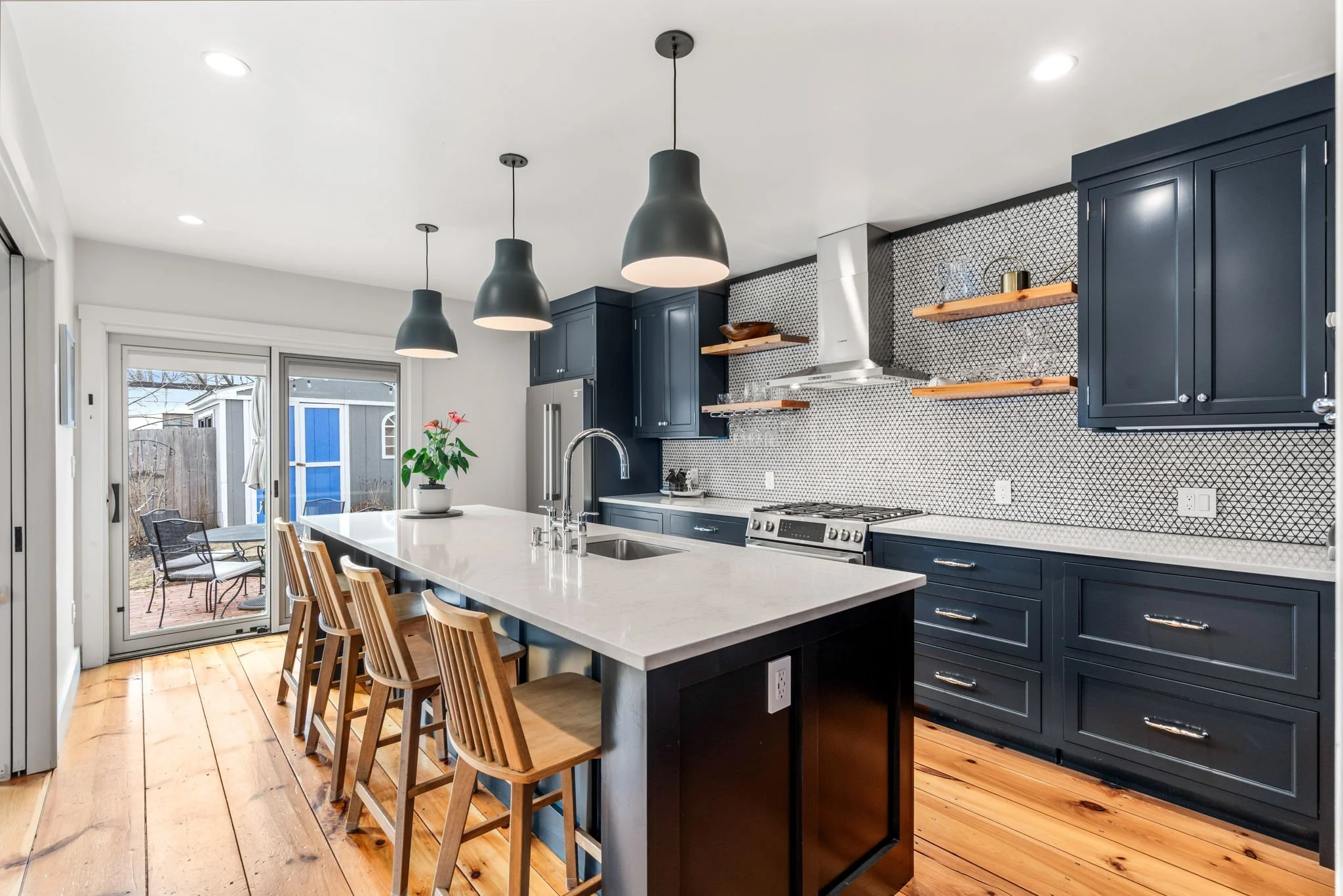 Modern kitchen with navy blue cabinets, white countertops, wooden bar stools, pendant lights, and sliding glass doors leading to an outdoor patio.