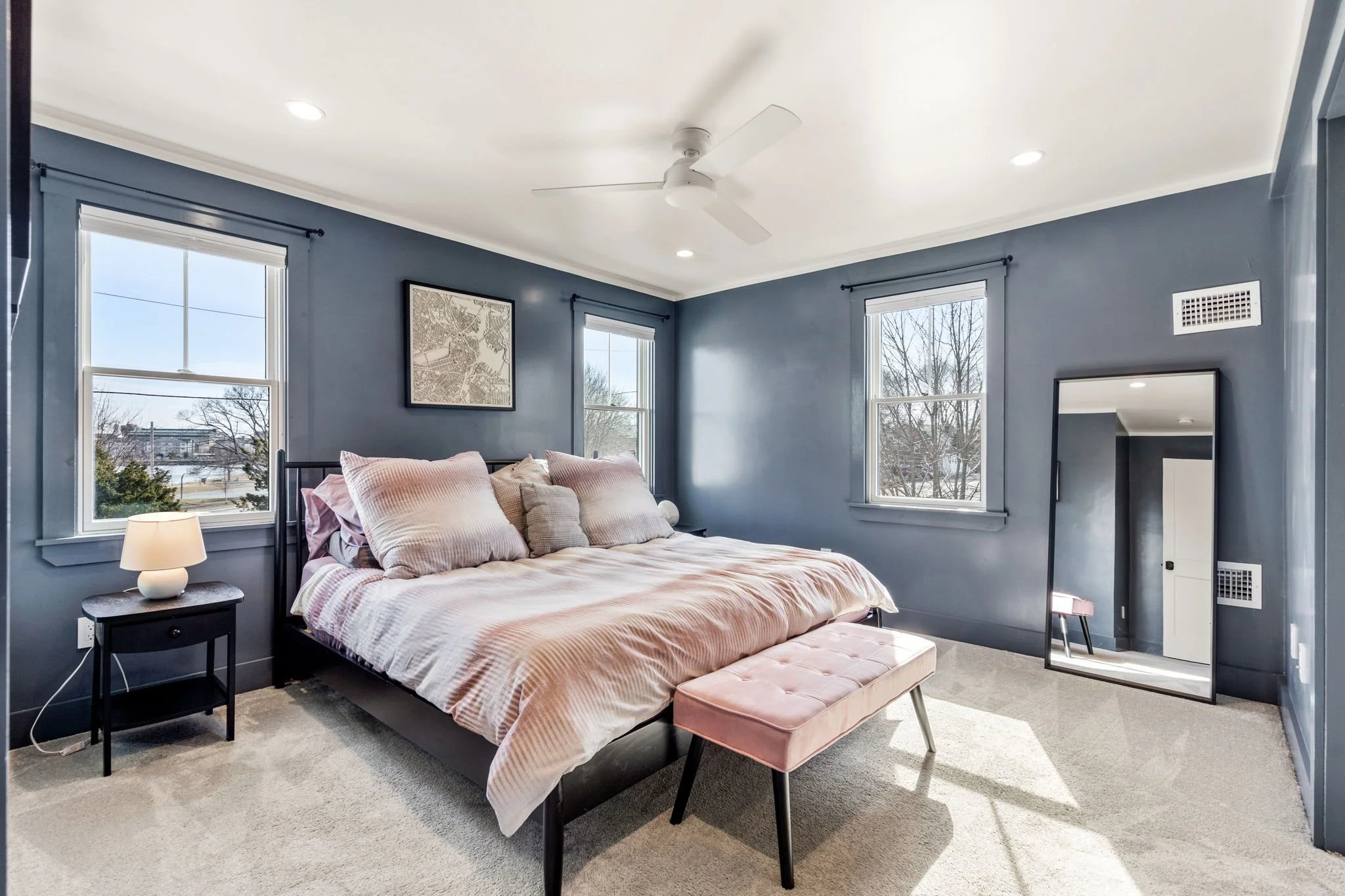 Bedroom with blue walls, three windows, a bed with pink and beige bedding, a pink tufted bench at the foot, a black nightstand with a lamp, a mirror leaning against the wall, and a ceiling fan.