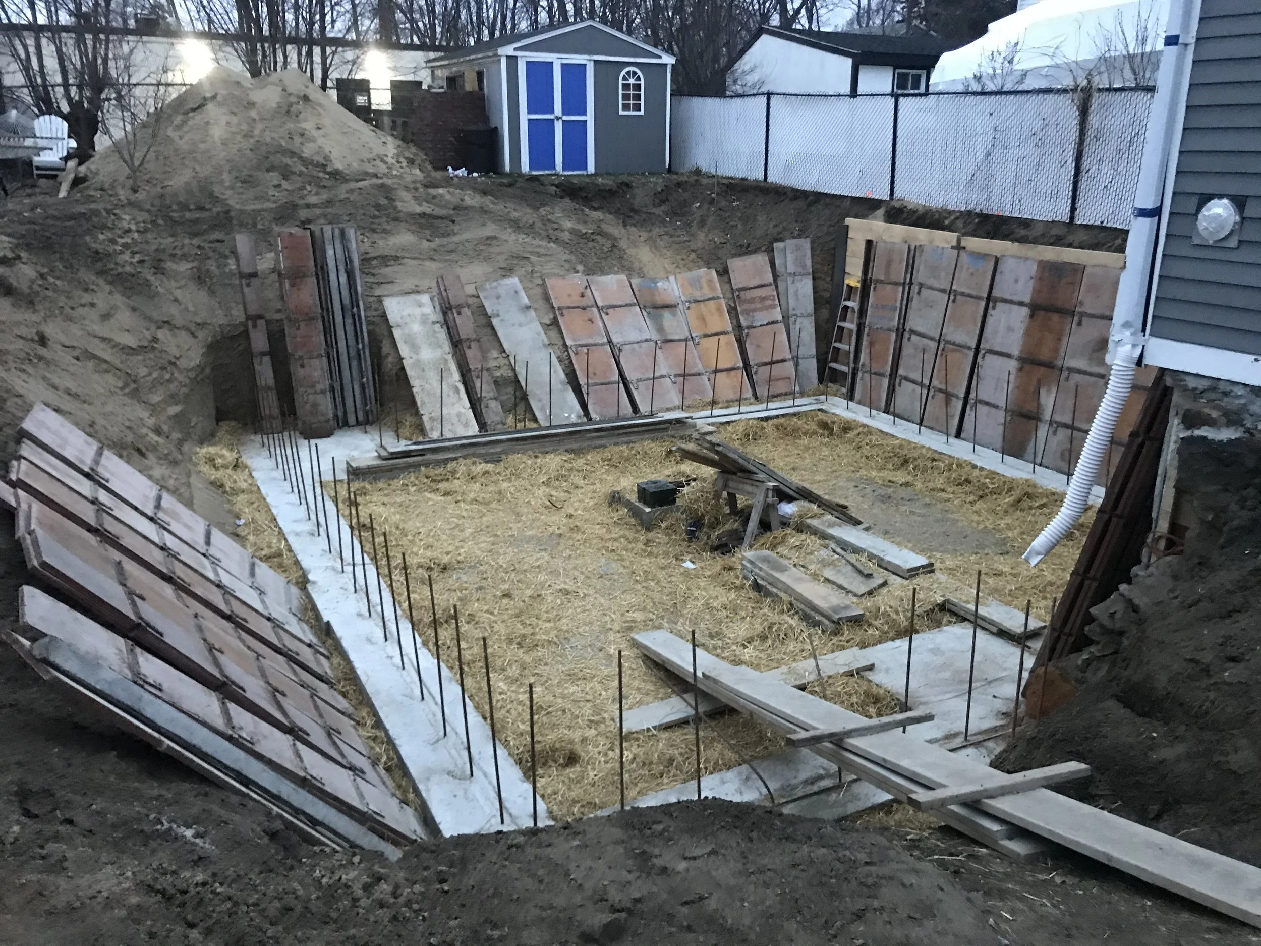 Construction site with foundation walls and rebar prepared for concrete pouring, wooden formwork in place, exposed soil, and construction tools, in a backyard.