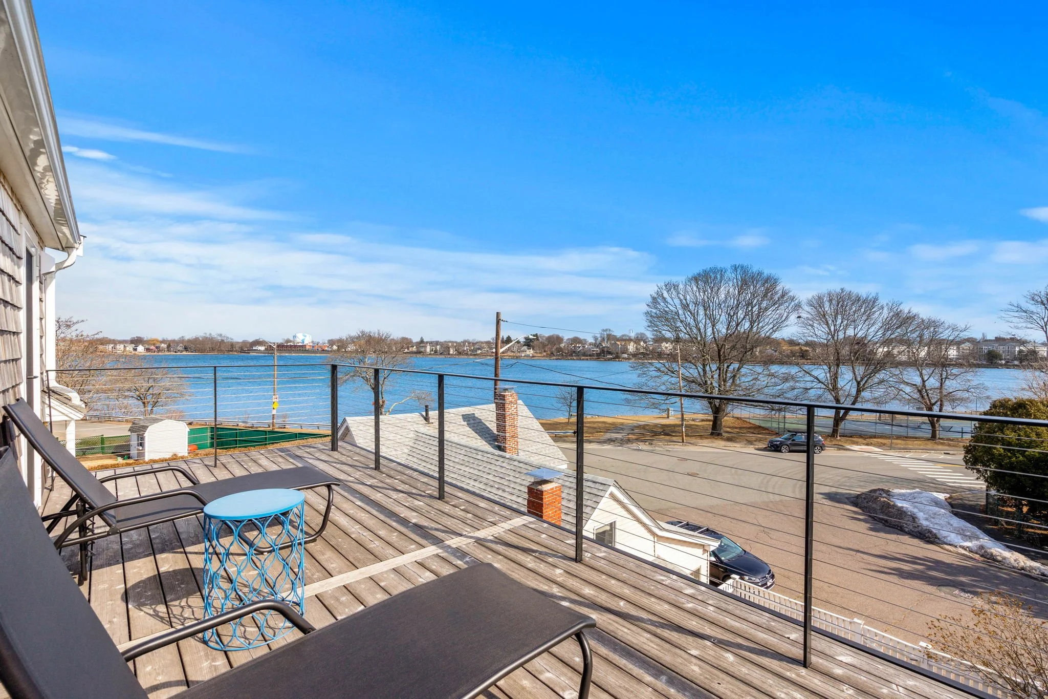 View of a lake from a wooden balcony with lounge chairs and a small blue table, trees without leaves, parked cars, and a partly cloudy blue sky.