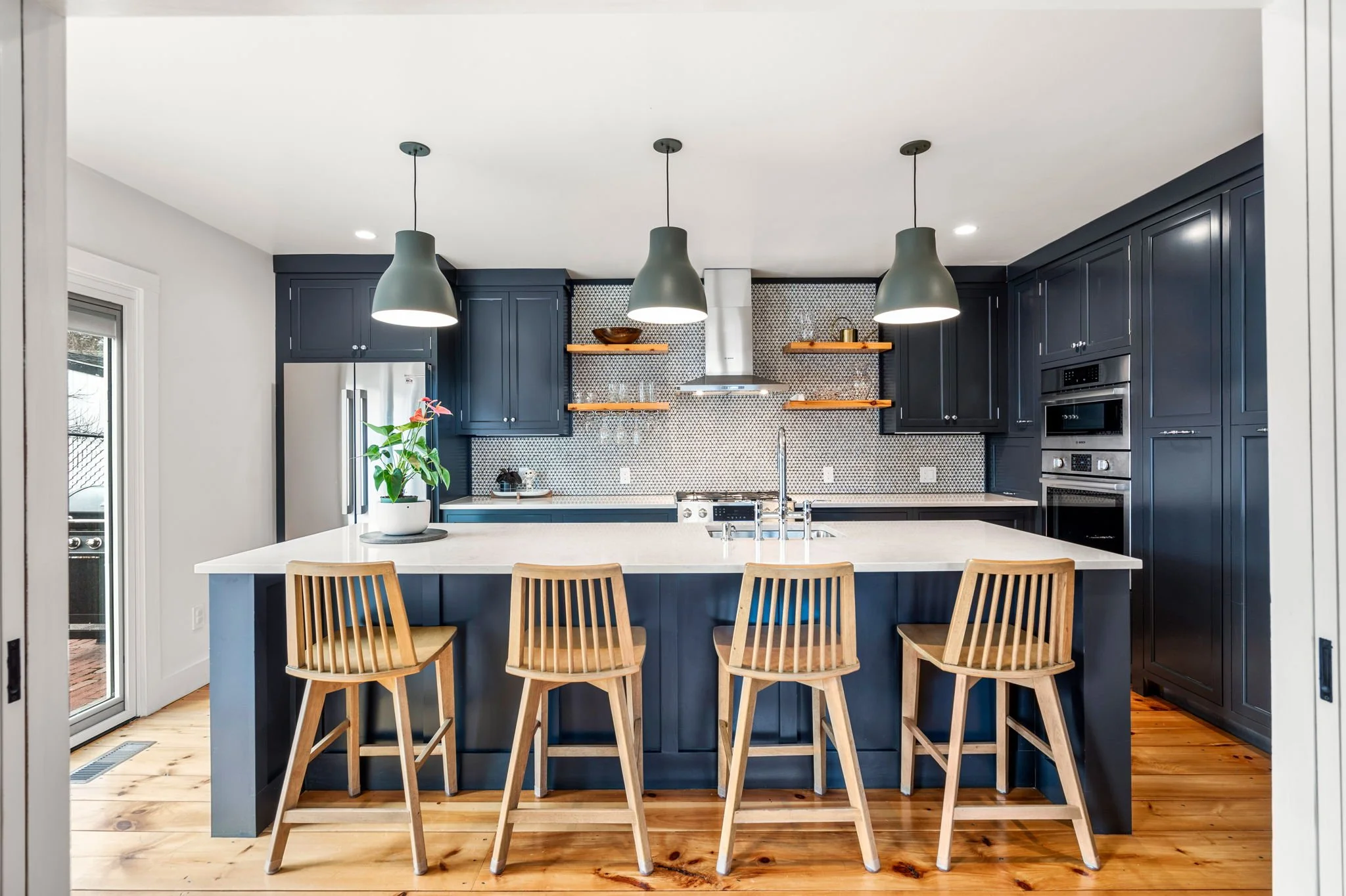 Modern kitchen with dark blue cabinets, white island, wooden barstools, stainless steel appliances, and pendant lighting.