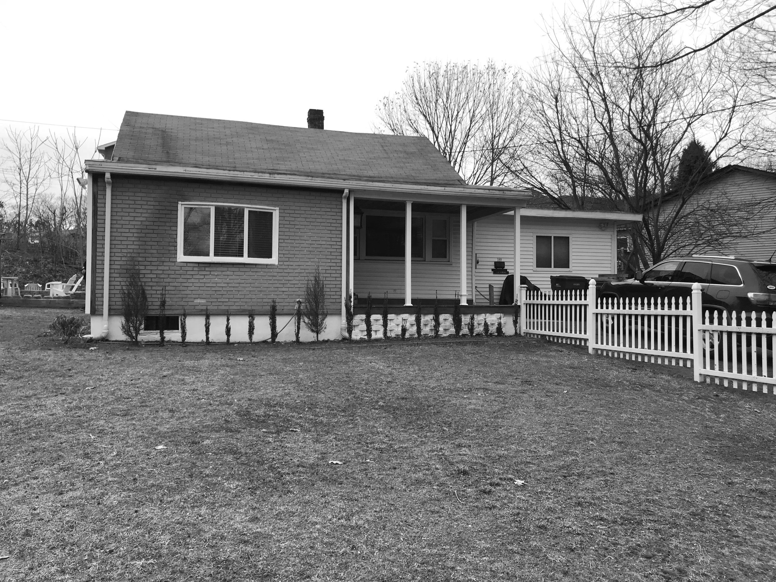 Black and white photo of a single-story house with a porch, white fence, and a parked car in the driveway. Bare trees are visible in the background.