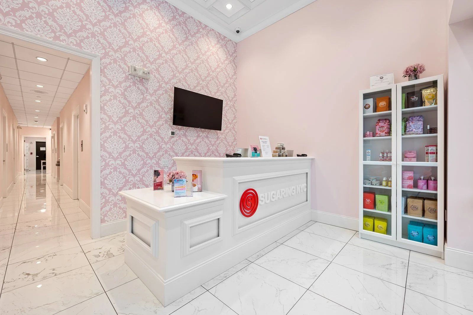 Pink reception area with a white counter that says 'SUGARING NYC', a black TV on the wall, and a white cabinet with colorful products on the right. The floor is marble, with a pink damask wallpaper on one wall and plain pink on the adjacent wall.