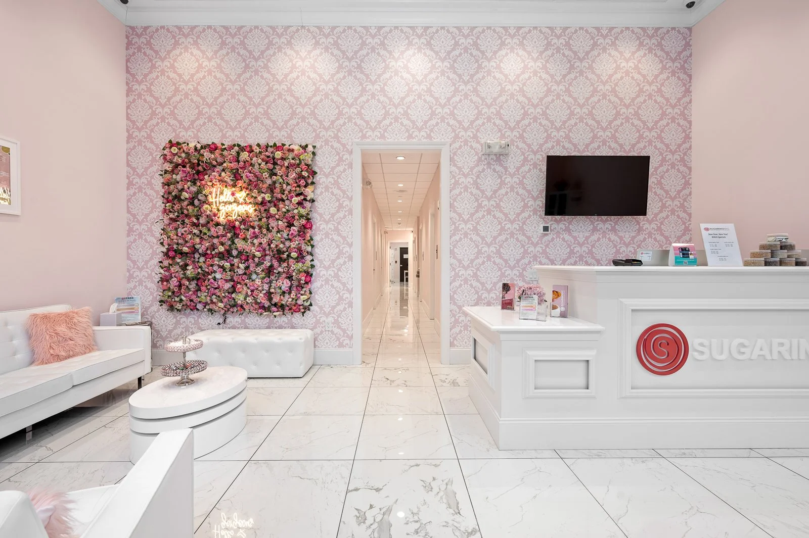 Interior of a pink-themed lounge or reception area with a white marble floor, pink damask wallpaper, a floral wall art with 'Hello Gorgeous' sign, white seating, a white counter with a Sugarfina logo, a wall-mounted TV, and a hallway in the backgroun