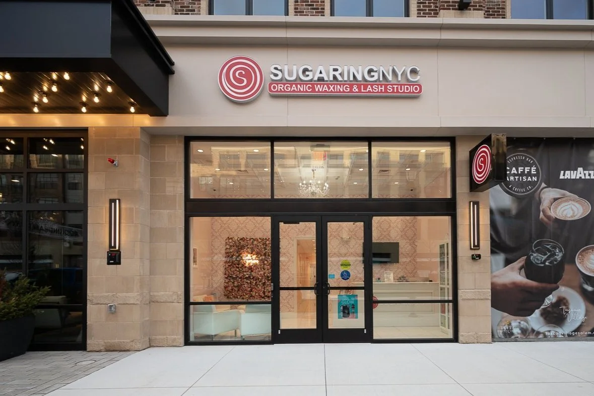 Exterior view of a business called SUGARING NYC, a waxing and lash studio in New York City, with large glass doors and signage.