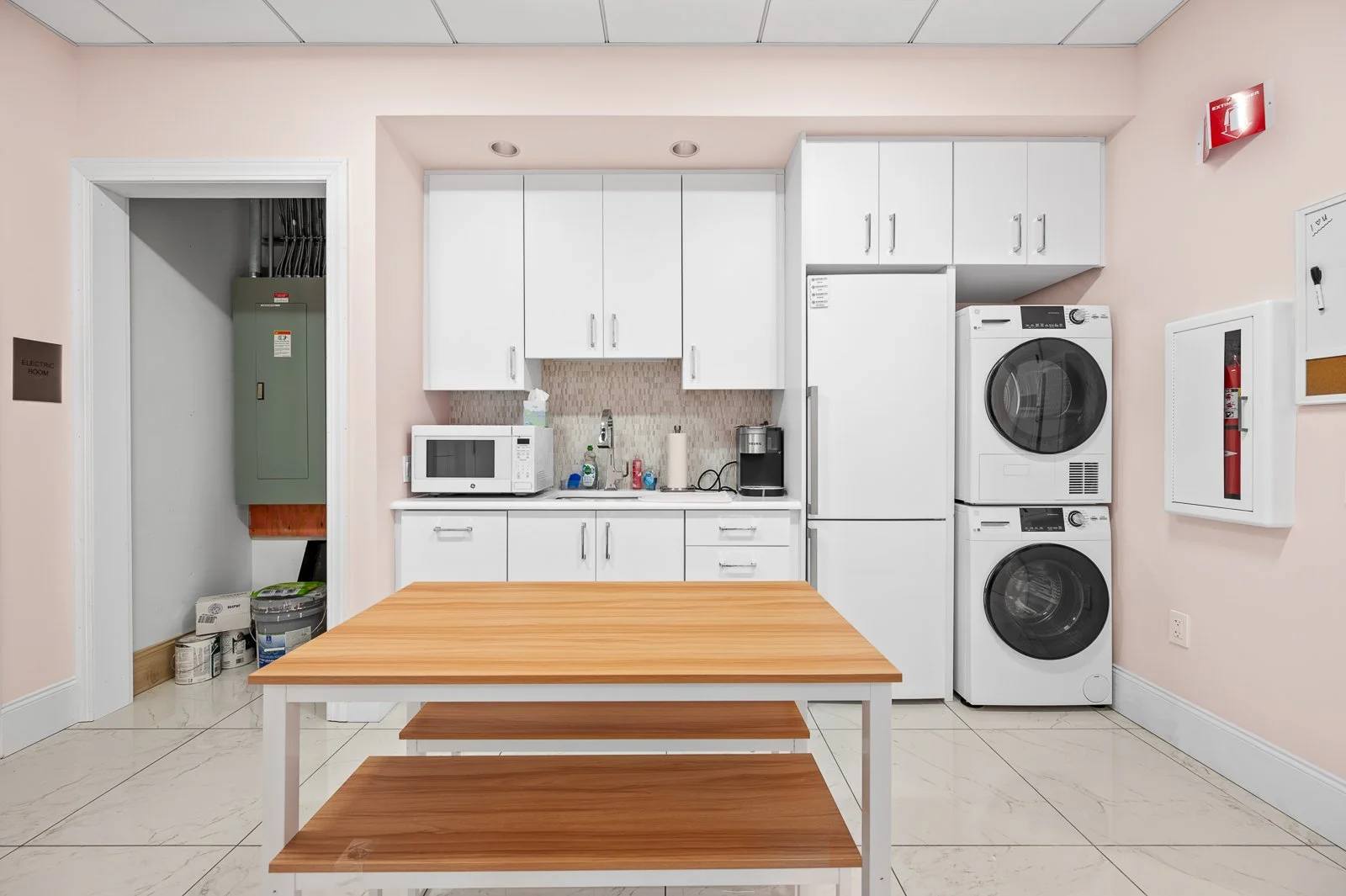 A laundry room with white cabinets, a microwave, sink, coffee maker, refrigerator, stacked washer and dryer, and a wooden table.
