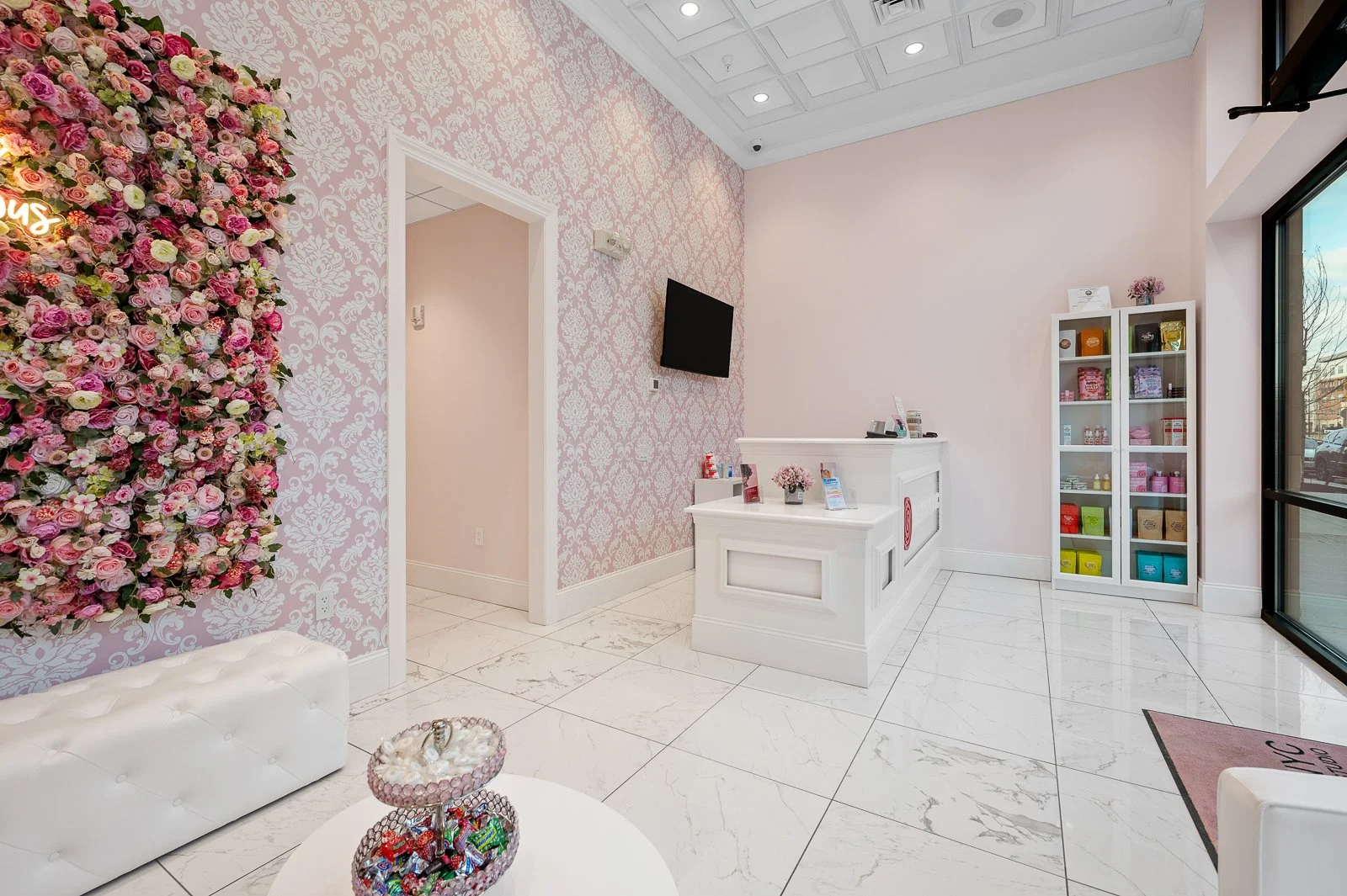 Interior of a brightly lit retail or beauty salon with pink and white decor, including a floral wall, a white reception desk, a glass display cabinet, and a large window showing the outside street.