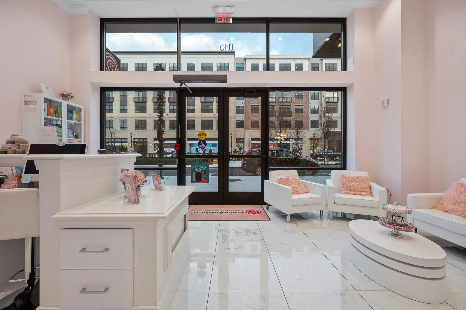 The interior of a modern waiting area with white furniture, pink pillows, and a reception desk, facing large glass doors and windows showing an urban street outside.