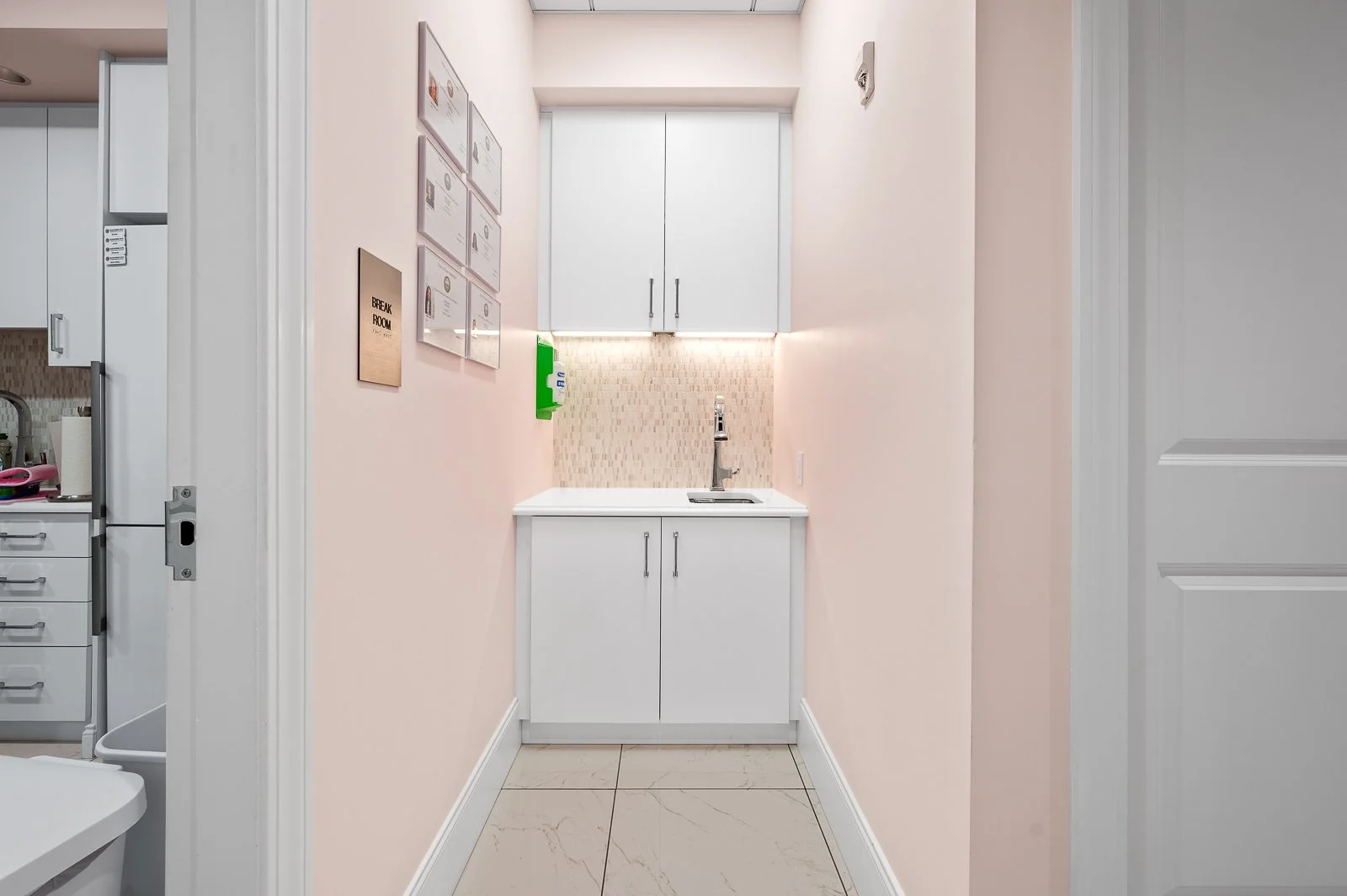 Small kitchenette with white cabinets, a beige tiled backsplash, a sink, and a wall sign that reads "Break Room."