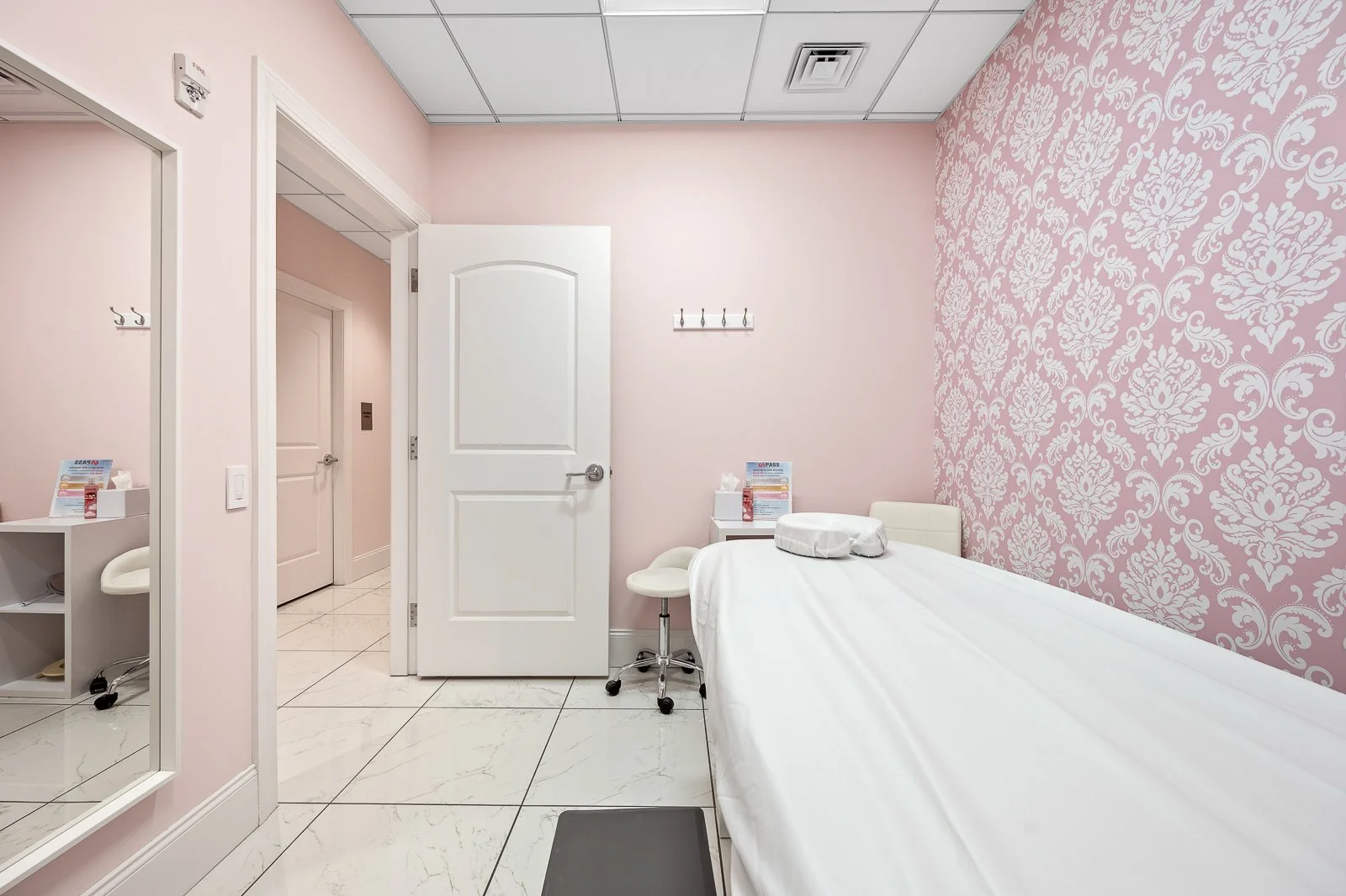 A medical examination room with a pink patterned accent wall, a white bed with a pillow, bedside table with tissues, a stool, and hooks on the wall.
