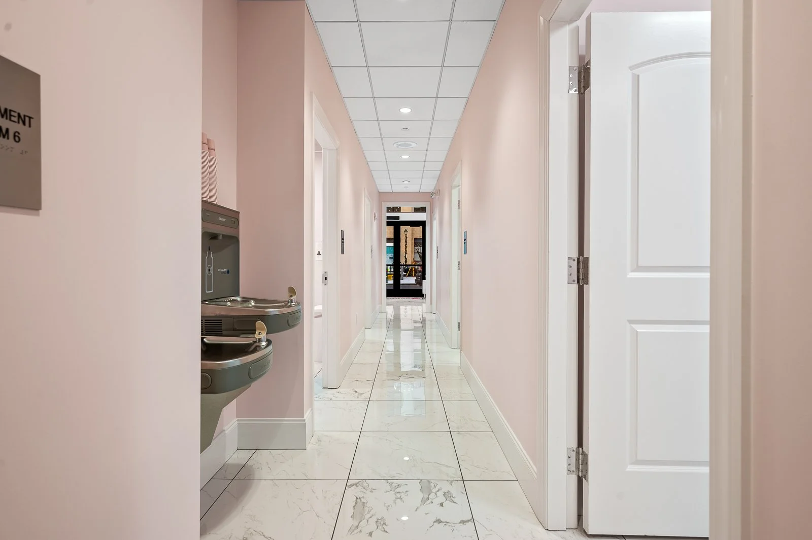 Empty hospital or clinic corridor with pink walls, white marble floor, and multiple rooms, one with an open door. Water fountain on the left side and a glass door at the end of the hallway.