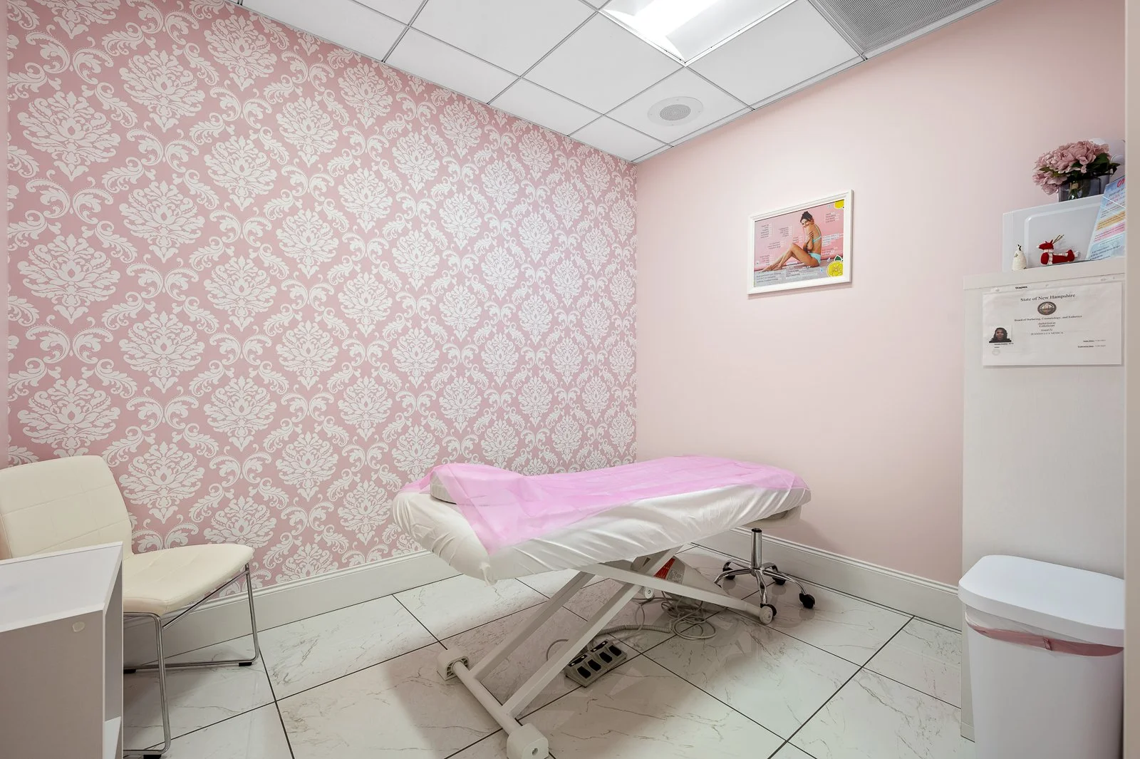 Medical examination room with pink and white colors, an examination table covered with pink paper, a white chair, a small white cabinet, framed picture on the wall, and a white trash bin.