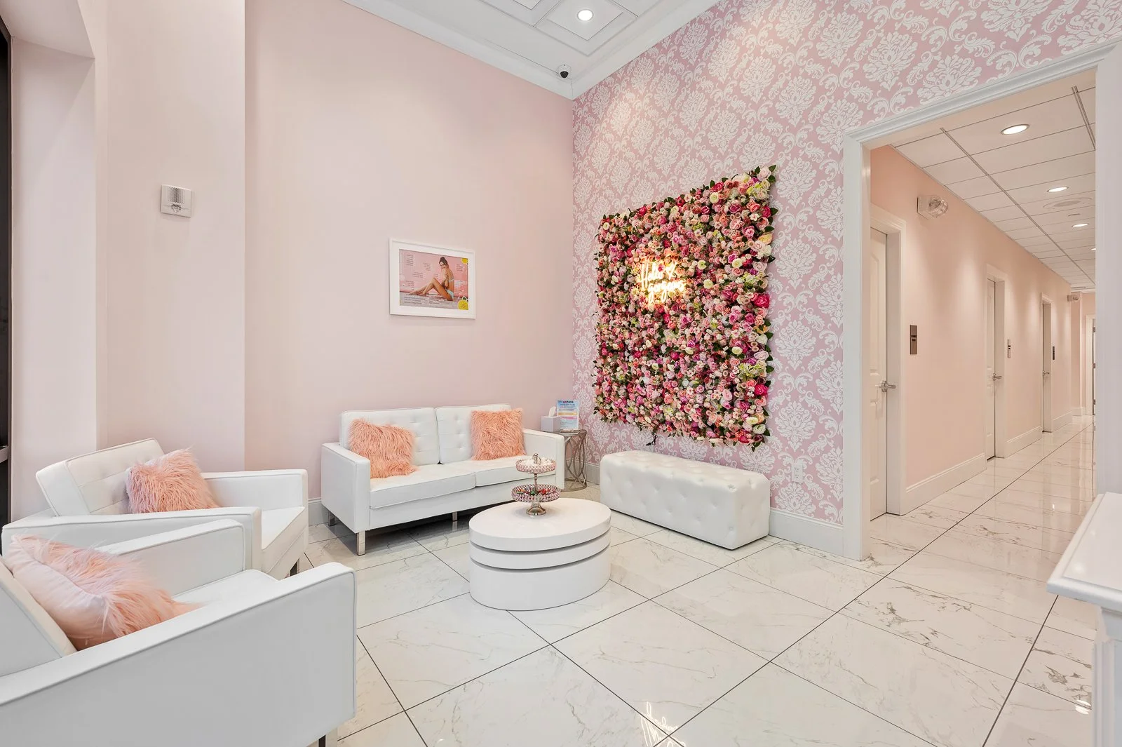 A lobby or waiting area with white sofas and pink fluffy pillows, pink wallpaper on one wall, a large floral art piece with pink and white flowers, and a small round coffee table at the center.