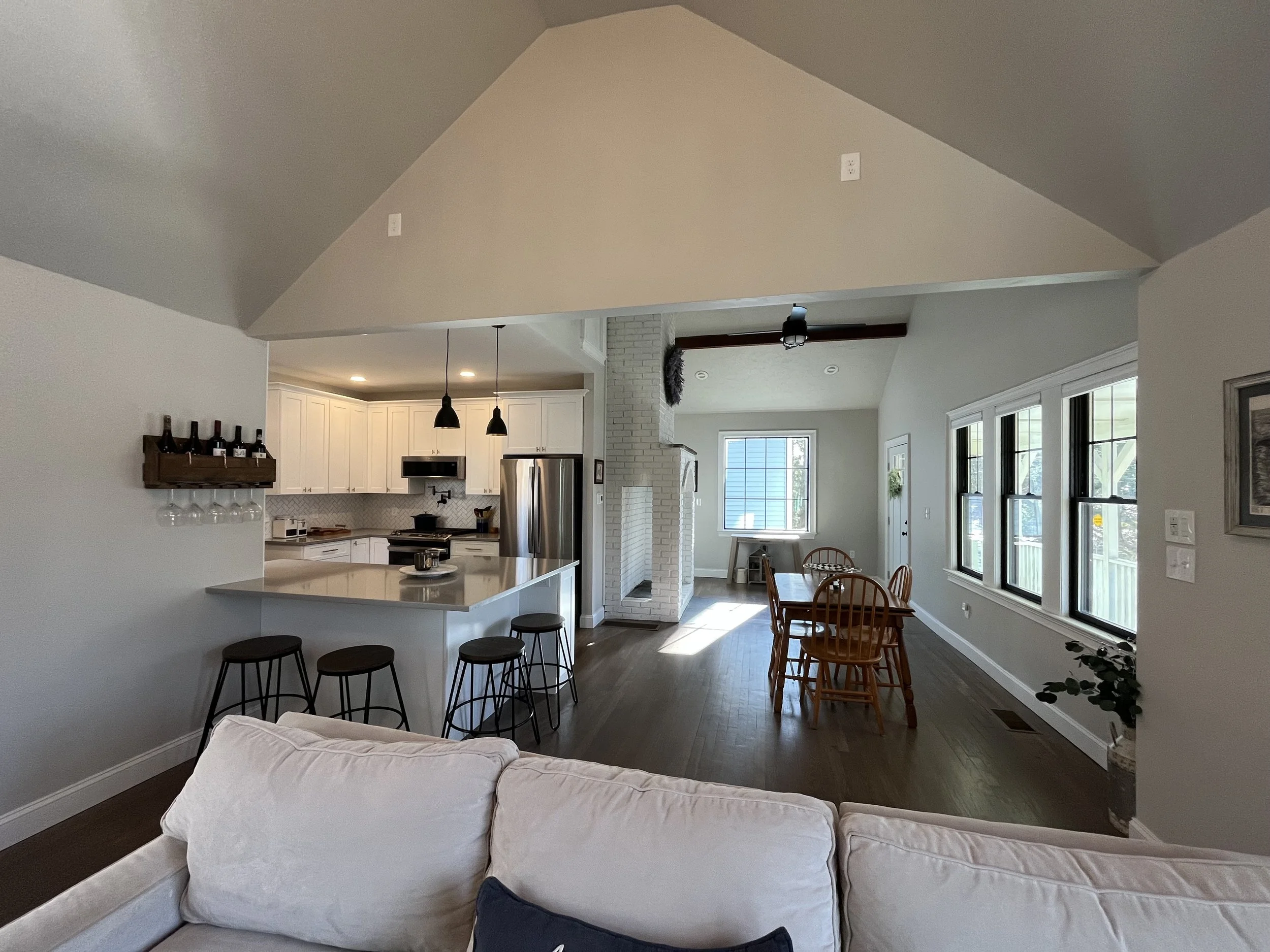 Open-concept living room, kitchen, and dining area with large windows, white cabinetry, stainless steel appliances, a kitchen island with bar stools, and a dining table with wooden chairs.