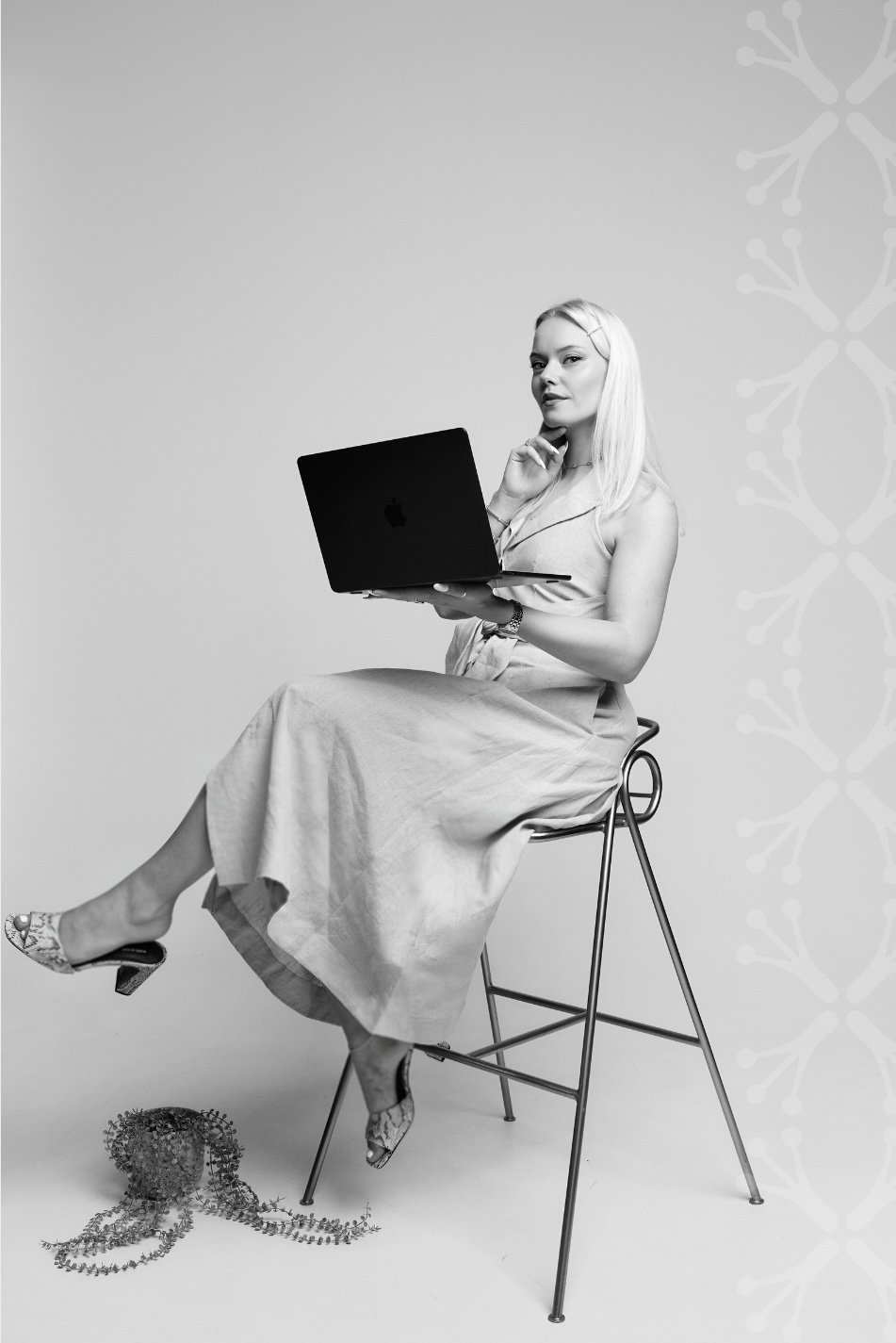 A black-and-white photo of Larissa sitting on a high stool with her legs crossed, holding a laptop. She has long blonde hair and is wearing a sleeveless dress and patterned high heels The background is plain with a subtle pattern on the right side.