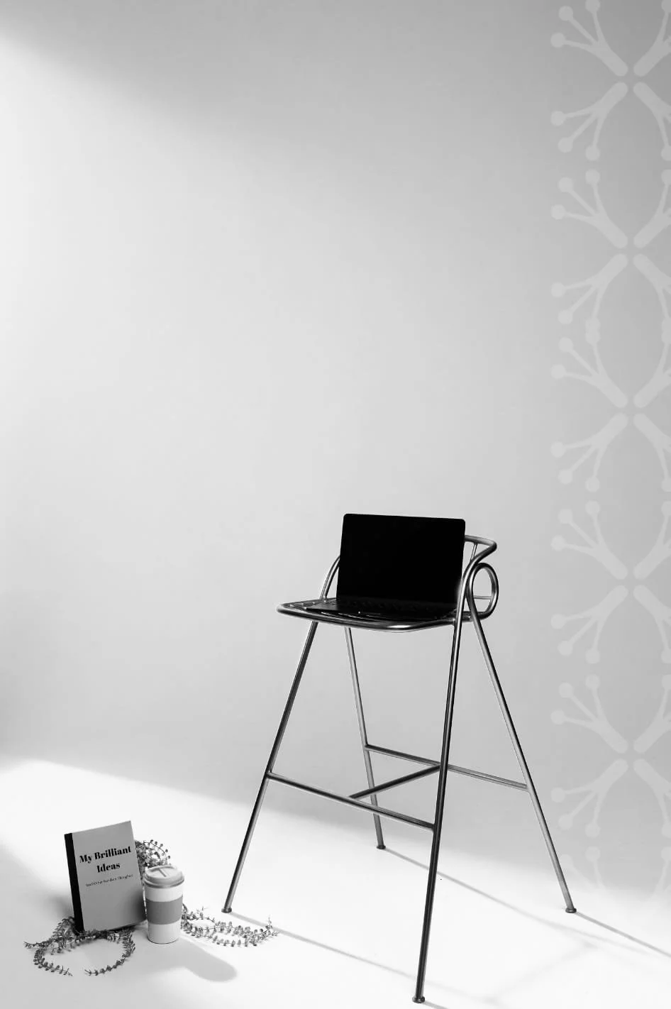 A black laptop on a metal stand with a book, a tumbler, and some jewelry on the floor nearby, set against a light gray and white background with a subtle pattern.