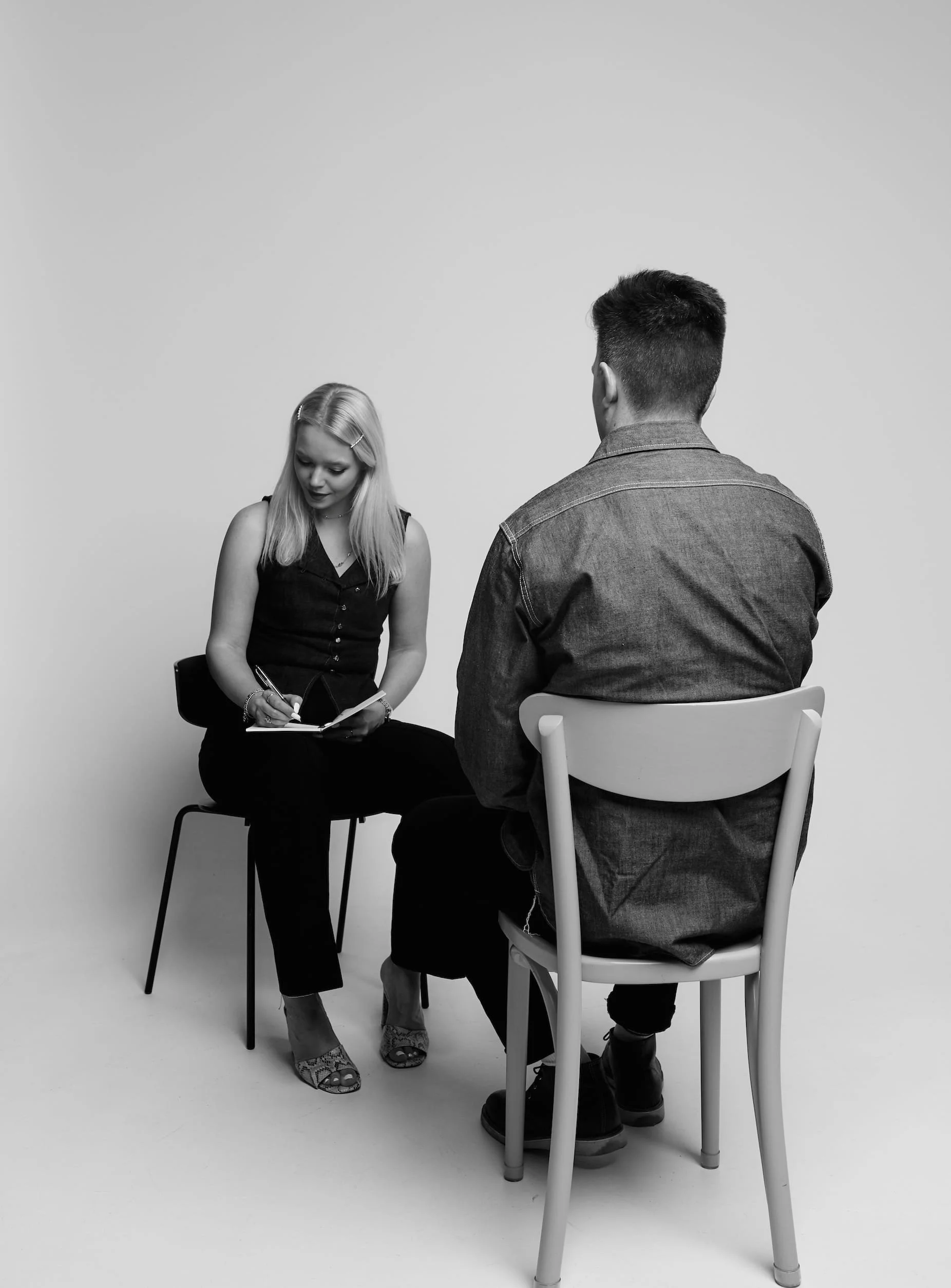 Black and white photo of Larissa with light hair and a man with dark hair sitting on chairs against a plain background. The woman is writing in a notebook, looking down. The man is facing away from the camera, wearing a denim shirt and dark pants.