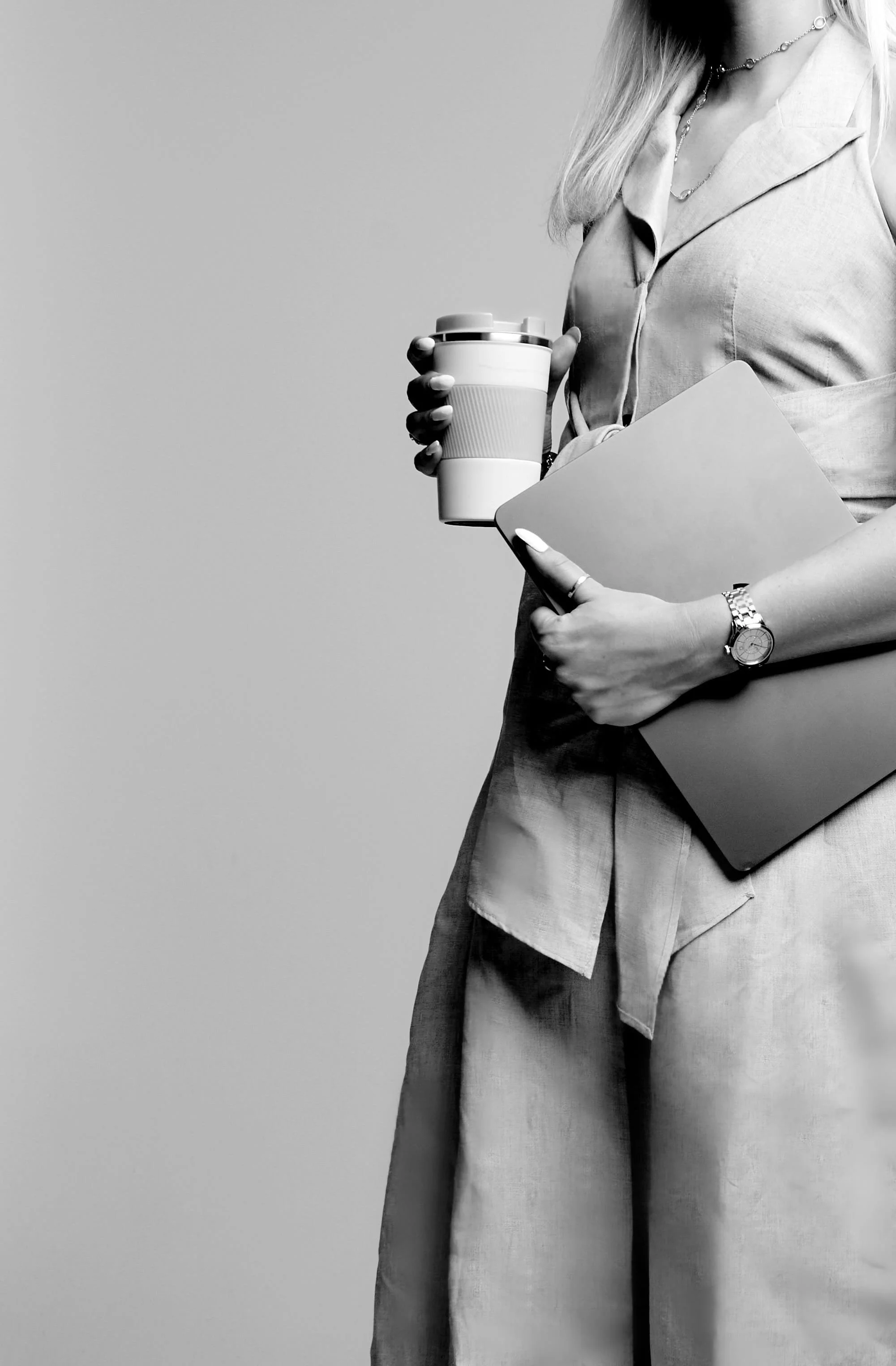 A woman, Larissa Hawkins, in business attire holding a coffee cup and a laptop, wearing a watch and jewellery, with a plain background.