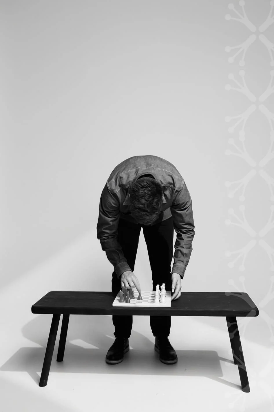 A man in a jacket playing chess on a small table in a studio with plain background.