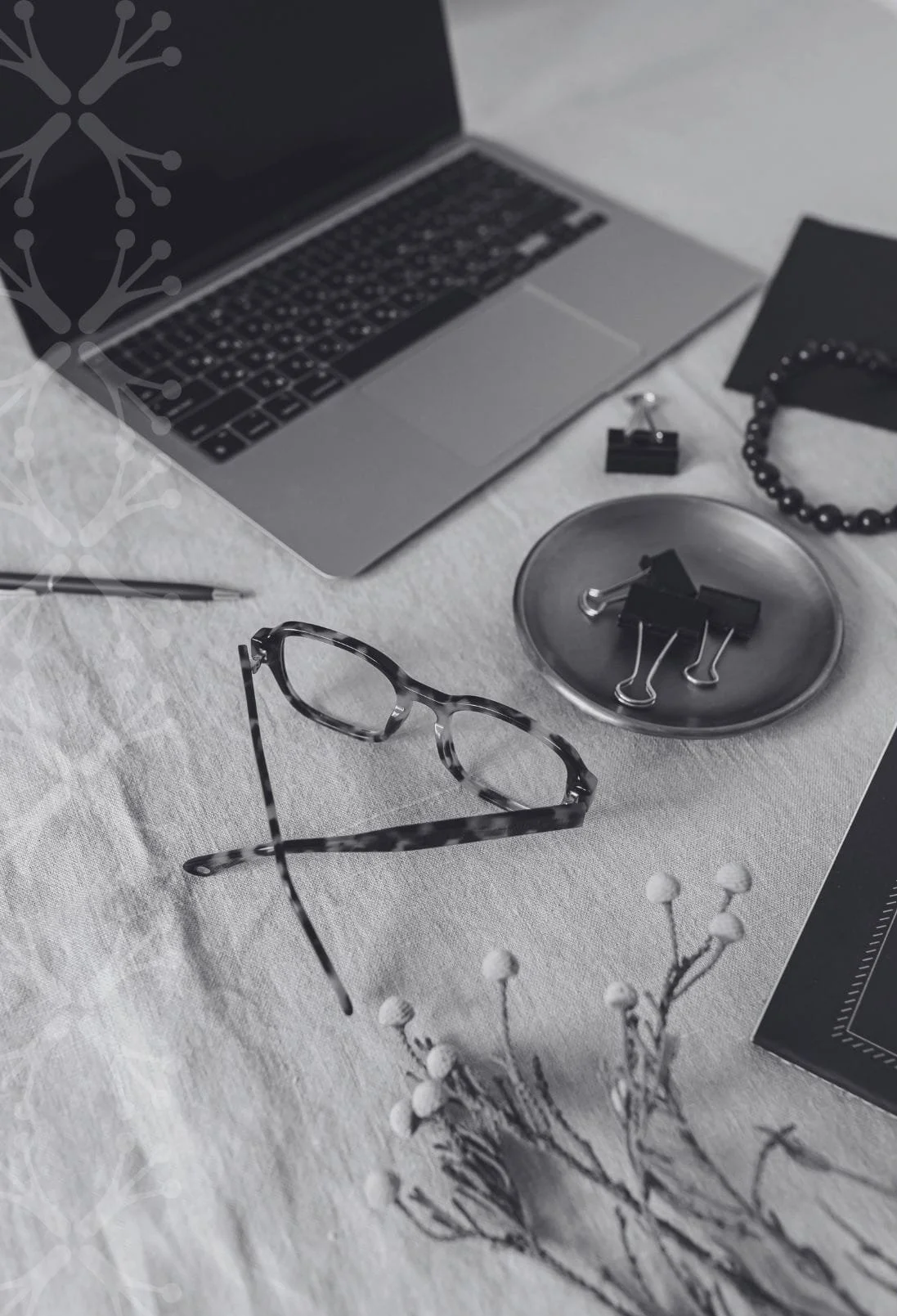 A desk with a laptop, pair of tortoiseshell glasses, a pen, a small dish with black binder clips, a black bead bracelet, a black notebook, and a sprig of white berries on a white textured surface.
