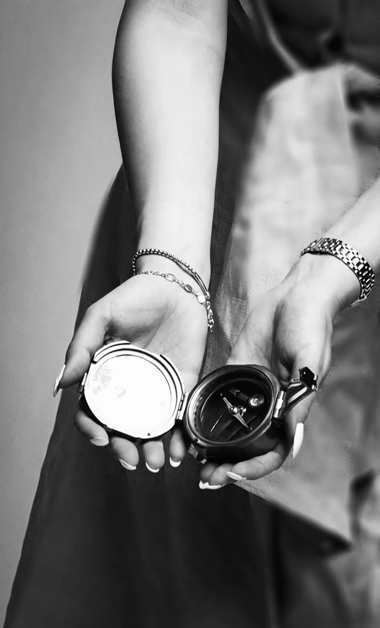 Black and white photo of a person's hands holding a wristwatch, with the watch face open, revealing the internal mechanism, surrounded by silver jewelry, with the person's arm and torso partially visible.